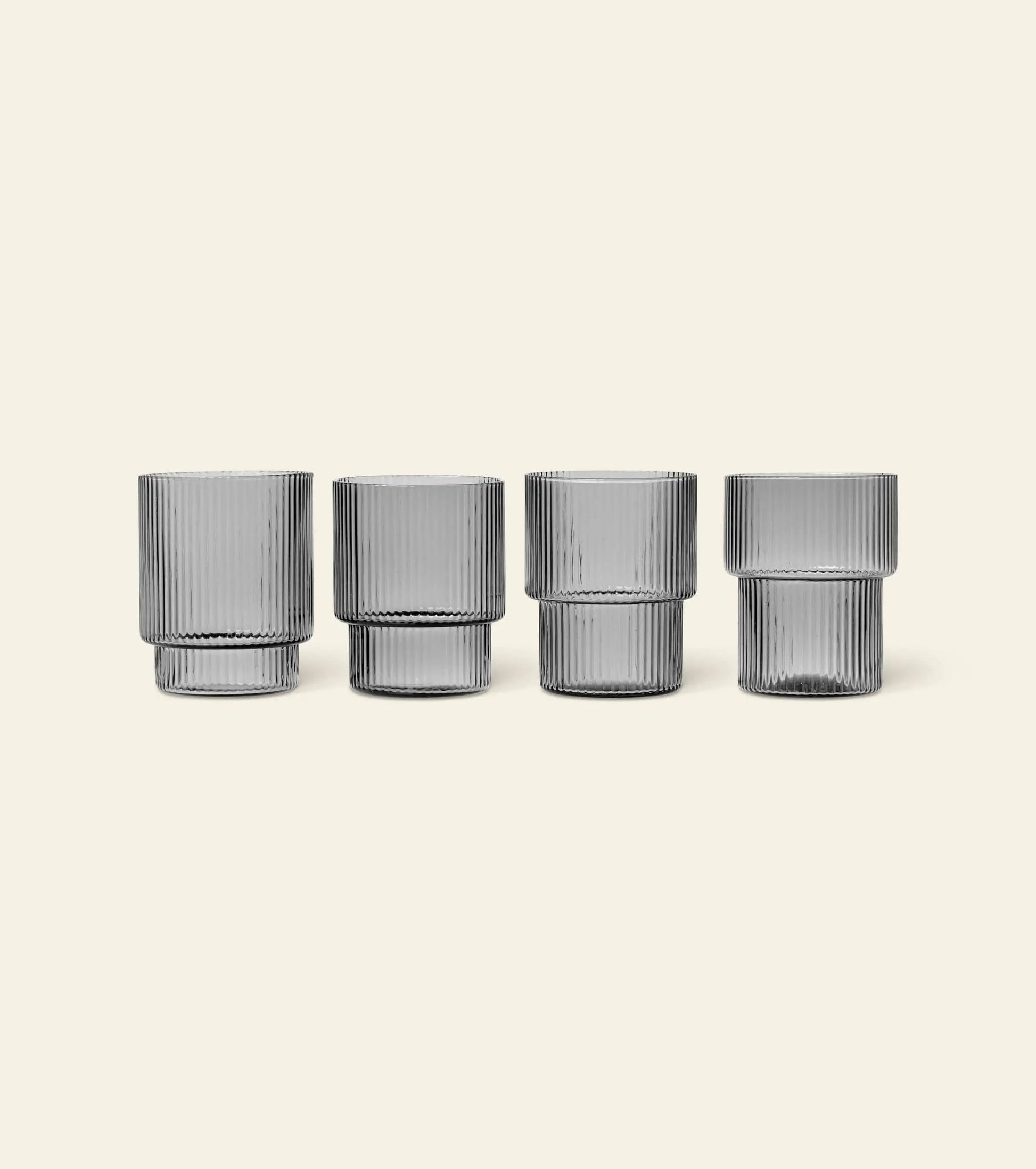 The ferm LIVING Ripple Drinking Glasses, Set of 4, features clear ribbed glasses with straight sides and a modern design from the brand ferm LIVING, shown in a row against a light beige background.