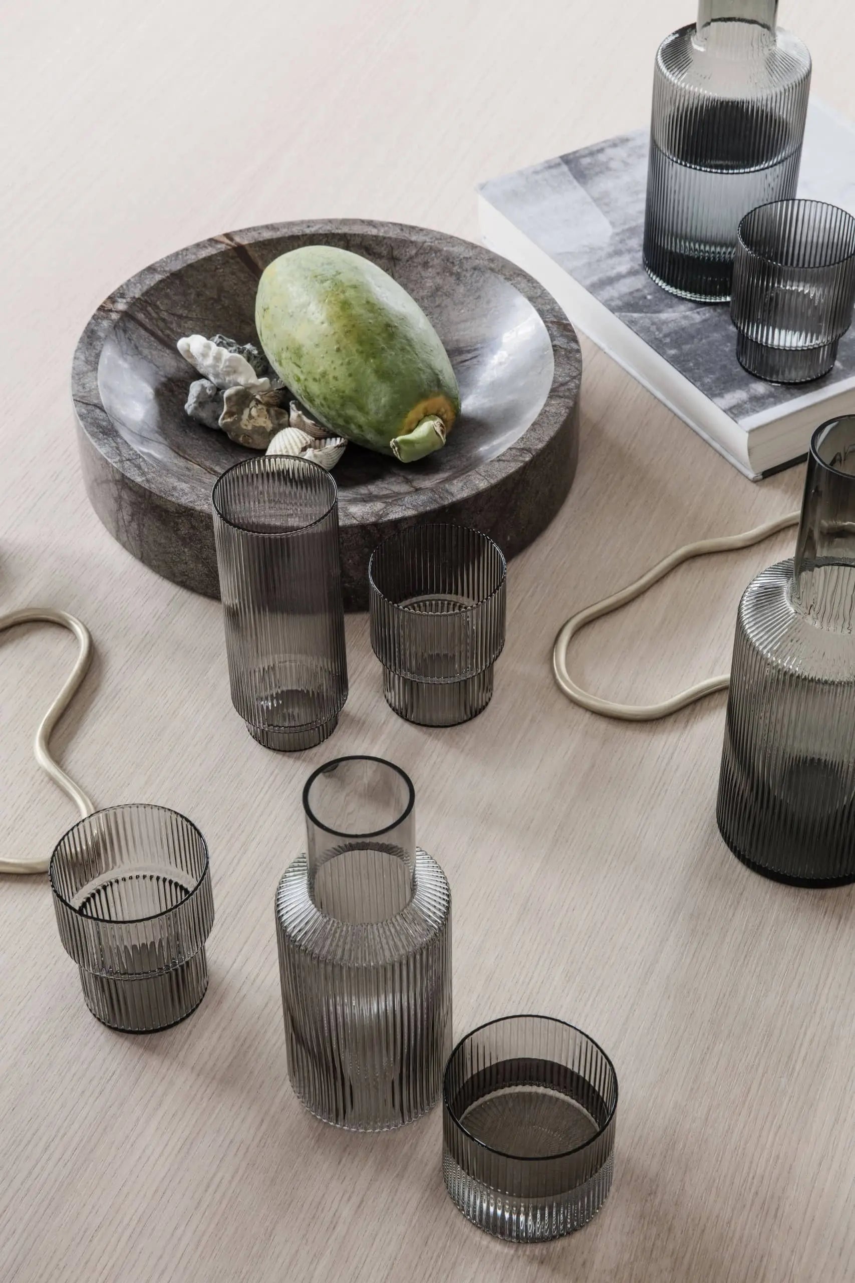 A wooden bowl with a green mango and shells sits beside the ferm LIVING Ripple Drinking Glasses, Set of 4, a book, and tan decorative ropes for a minimalist, modern look.