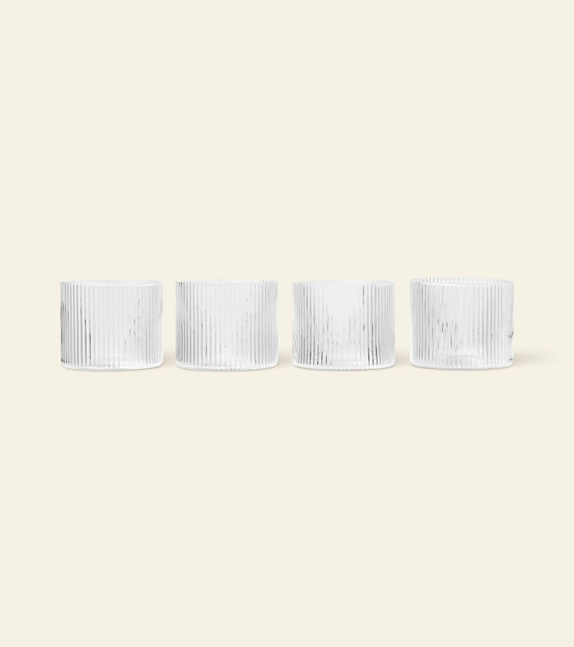 Four white, cylindrical ceramic cups with vertical ribbed lines are arranged in a row on a light beige background, mirroring the elegant design of the ferm LIVING Ripple Low Glasses, Set of 4 by ferm LIVING.