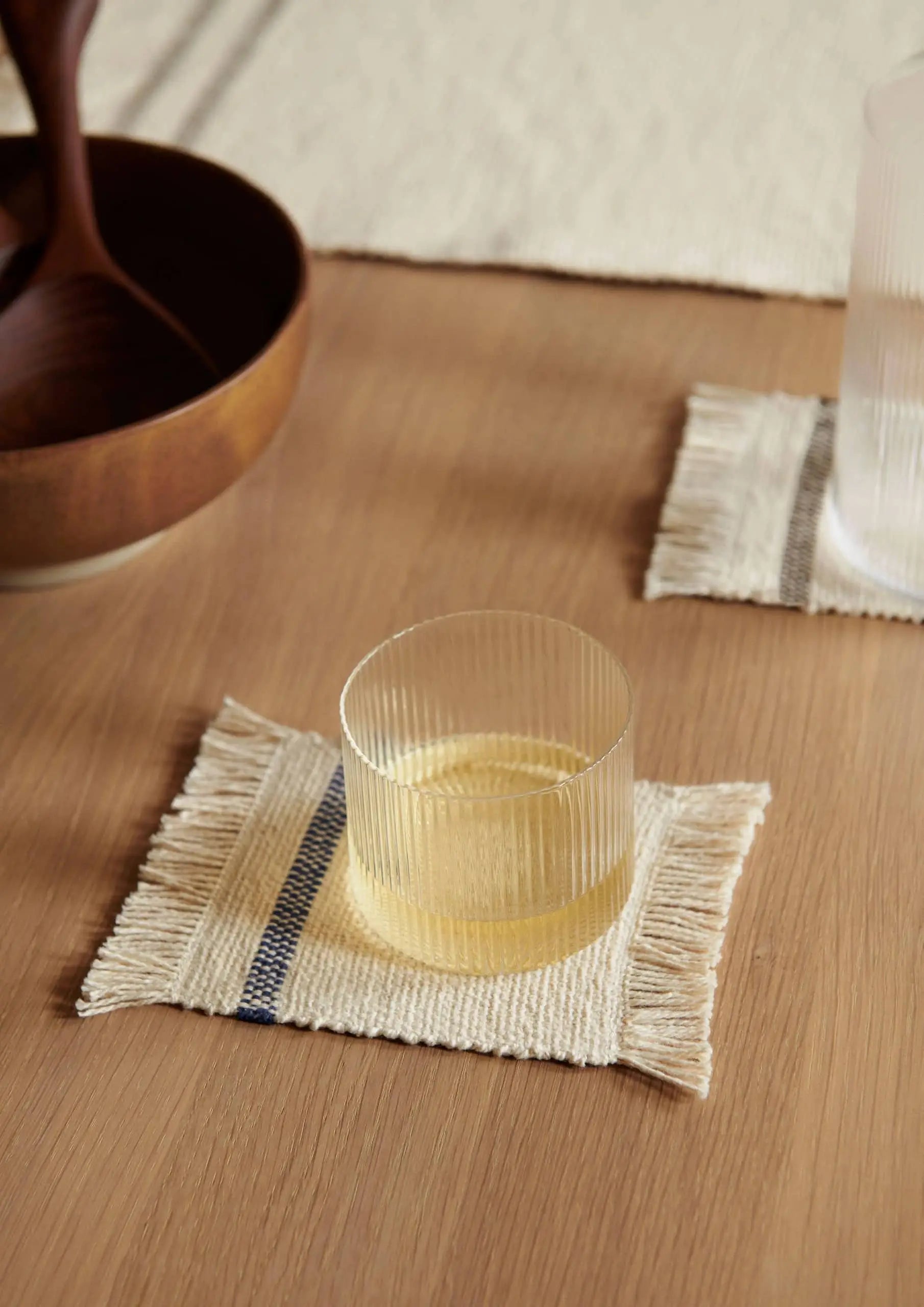 A ferm LIVING Ripple Low Glass from the set of 4 rests on a woven coaster atop a wooden table, accompanied by a wooden bowl and another matching cocktail glass, both also on coasters.