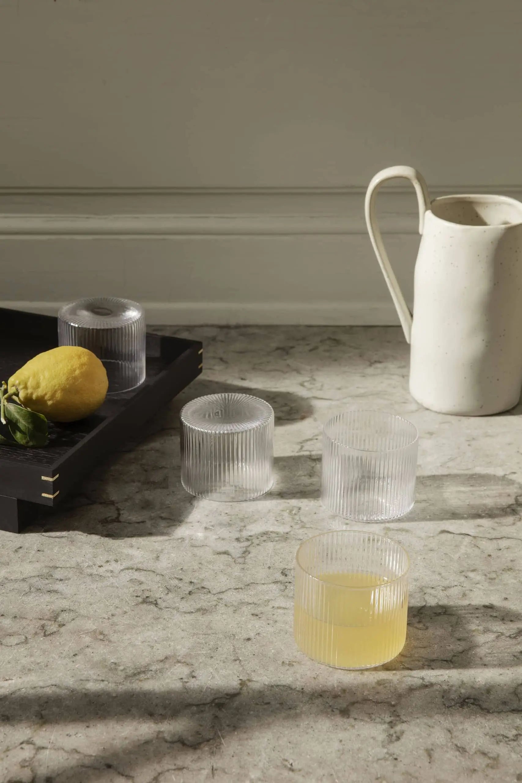 Four ferm LIVING Ripple Low Glasses, one with yellow juice, rest on a marble counter beside a white ceramic pitcher. A black tray holds a lemon and two empty cocktail glasses as sunlight creates gentle shadows.