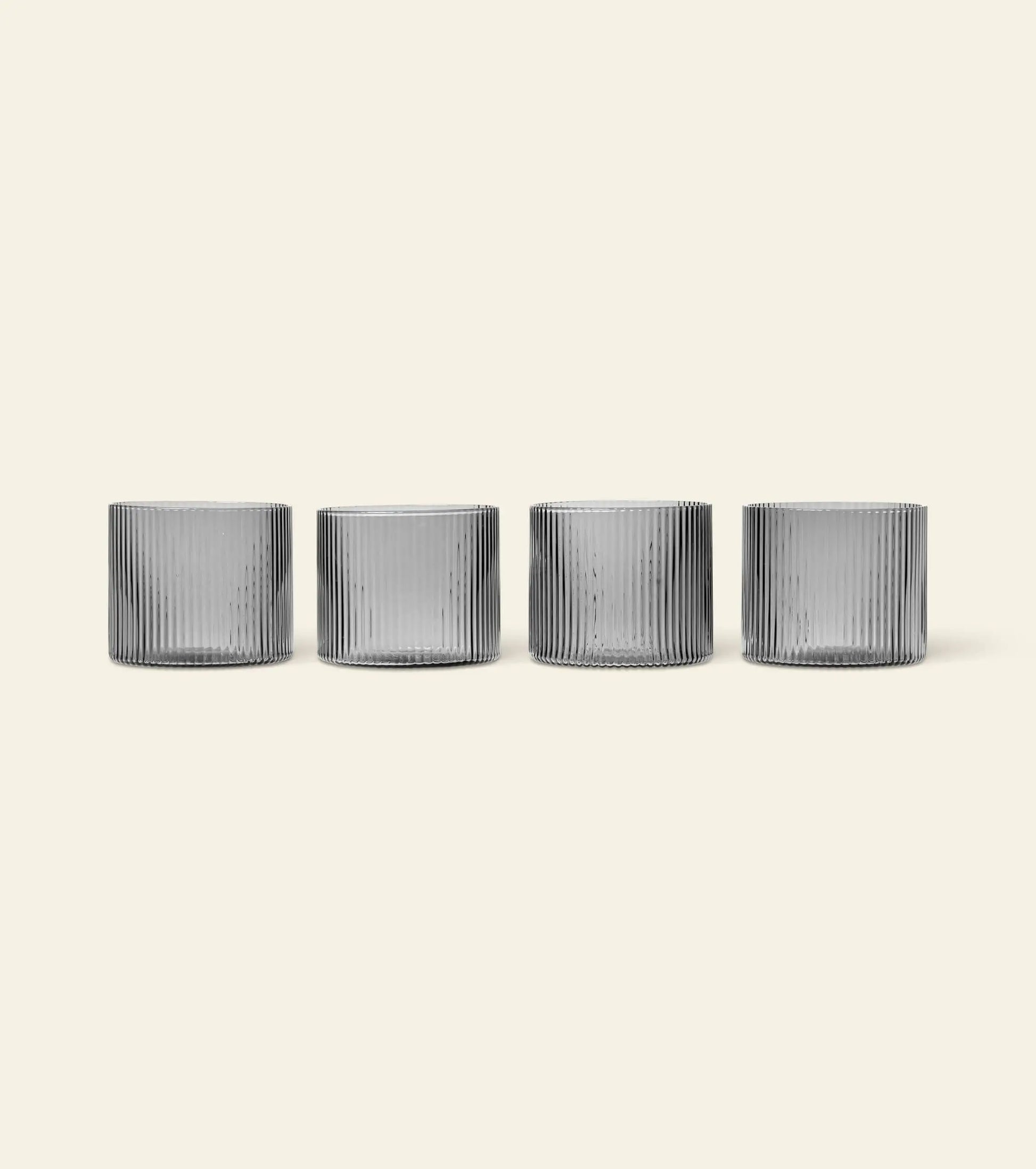 The ferm LIVING Ripple Low Glasses, Set of 4, feature vertical ridges and are stylishly arranged in a row on a light beige background—ideal for serving cocktails.