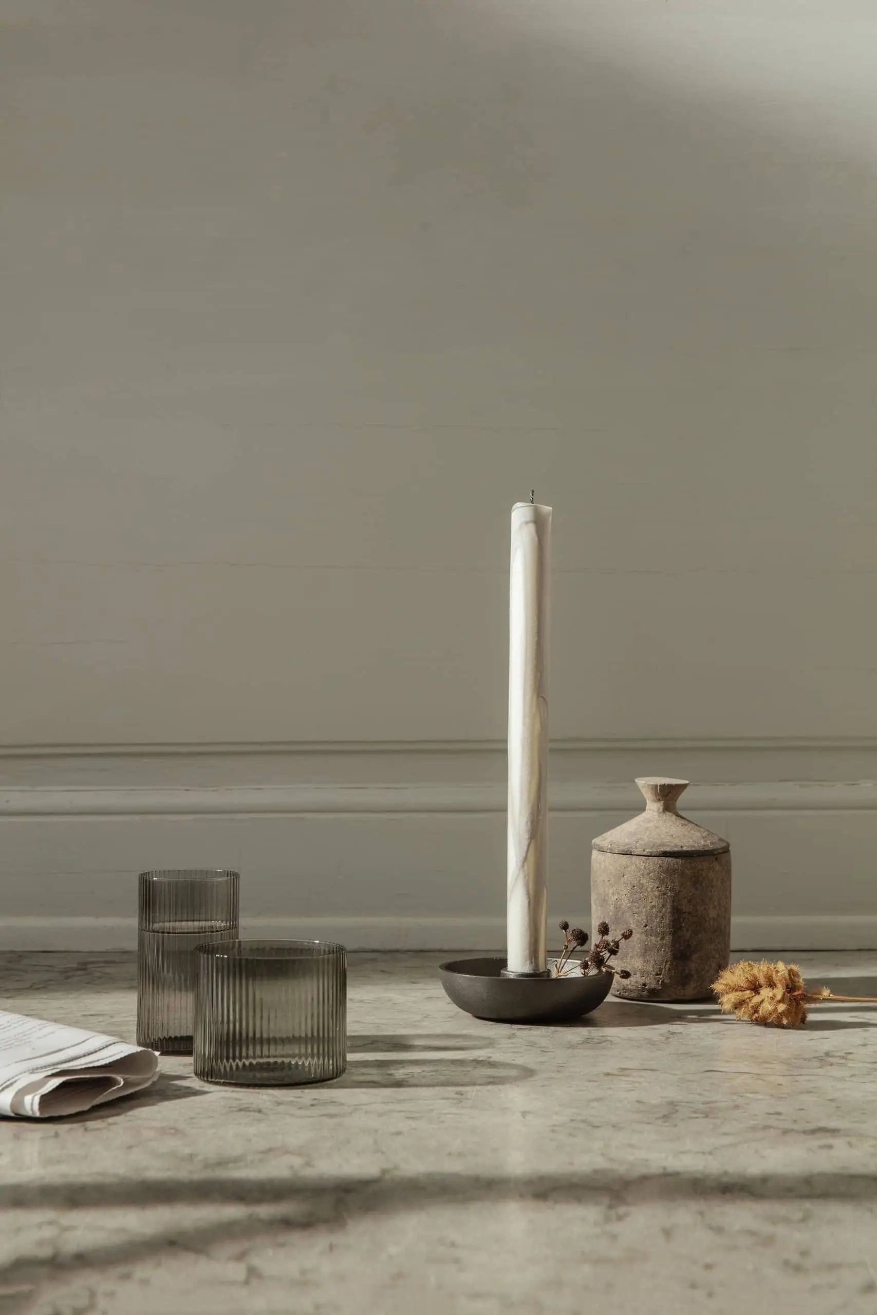 Minimalist scene with a tall white candle in a black holder, a rustic ceramic vase, dried flower, folded cloth, and two ferm LIVING Ripple Low Glasses on a light surface against a plain light wall.