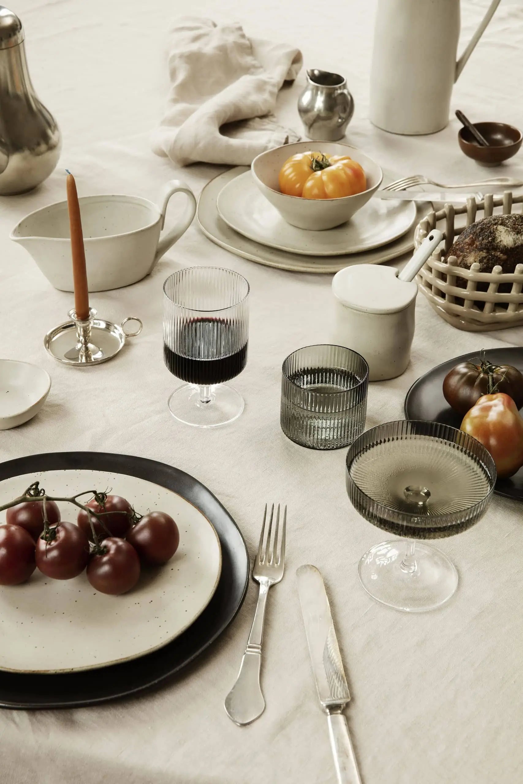 An elegant table setting features plates of tomatoes, ferm LIVING Ripple Low Glasses (set of 4) filled with drinks, a lit candle, cutlery, a ceramic pitcher, a bread basket, and neutral linens on a cream tablecloth.