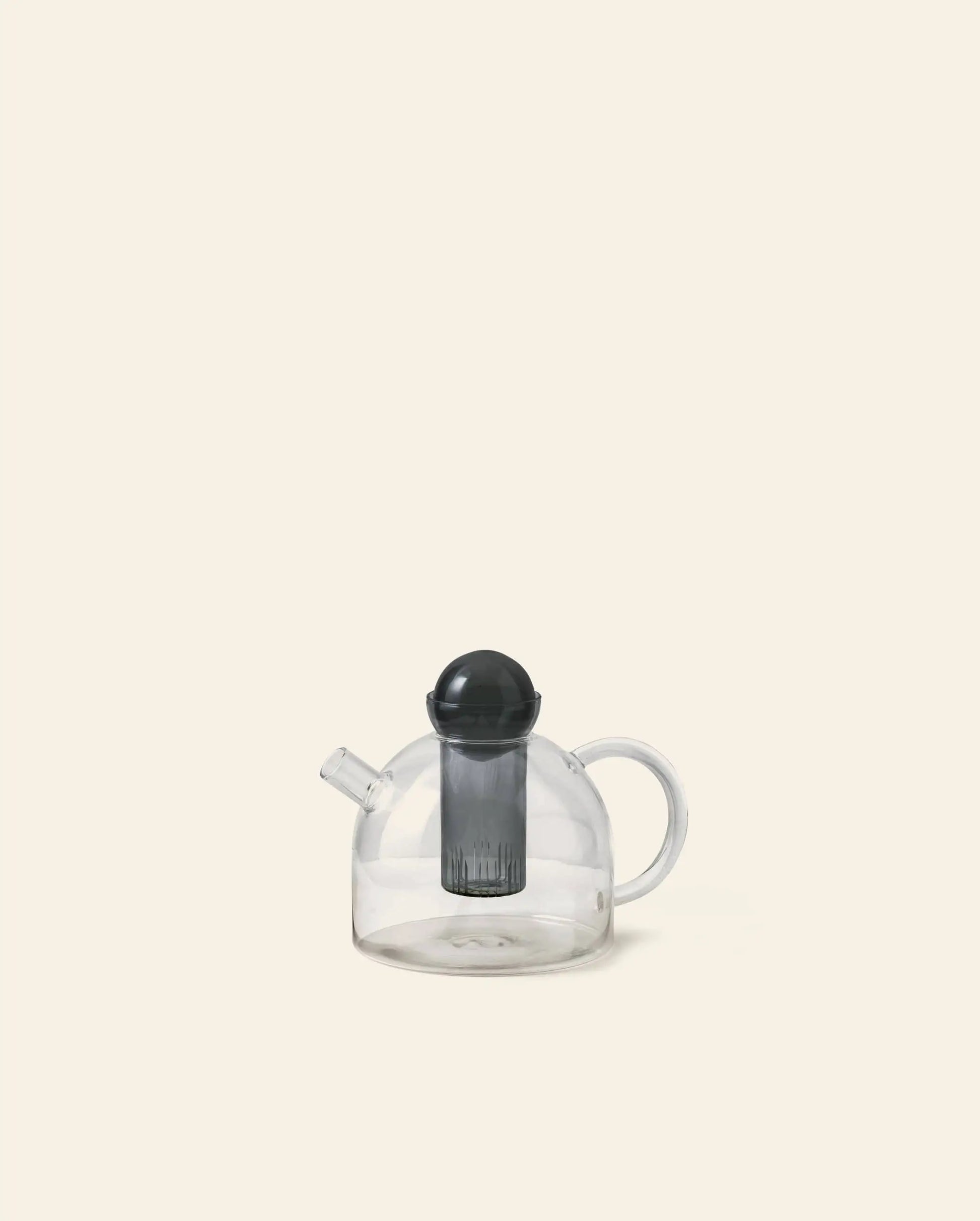The ferm LIVING Still Teapot by ferm LIVING is a clear, heat-resistant glass teapot with a round body, curved handle, short spout, and includes a black infuser and lid, shown against a plain beige background.