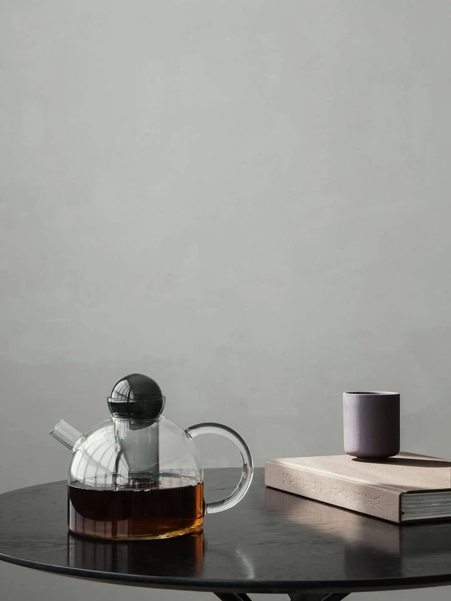The ferm LIVING Still Teapot by ferm LIVING, filled with dark tea, rests on a round black table next to a closed book and a small purple cup, set against a plain light gray background.