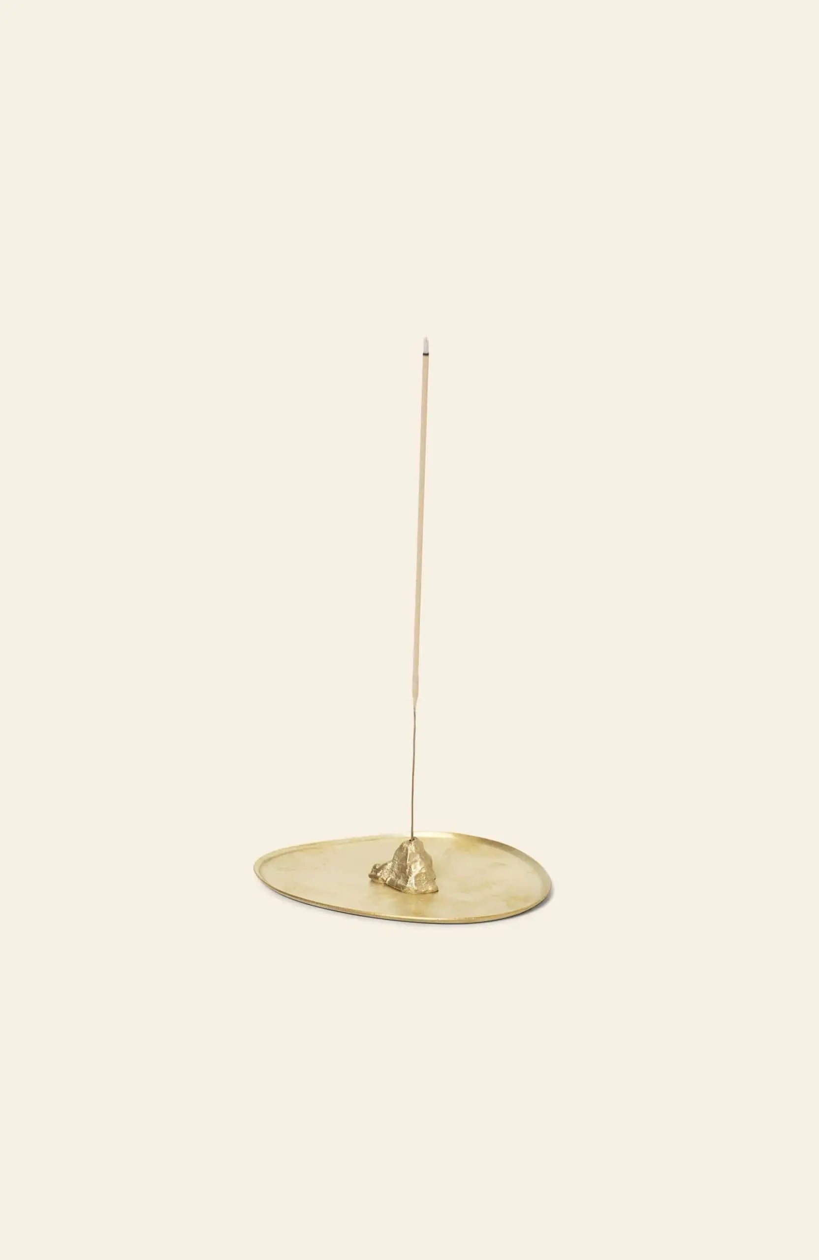 A thin incense stick stands upright in the ferm LIVING Stone Incense Burner, a minimalist gold holder resembling a small rock on an oval tray, adding serene Zen decor against a plain beige background.