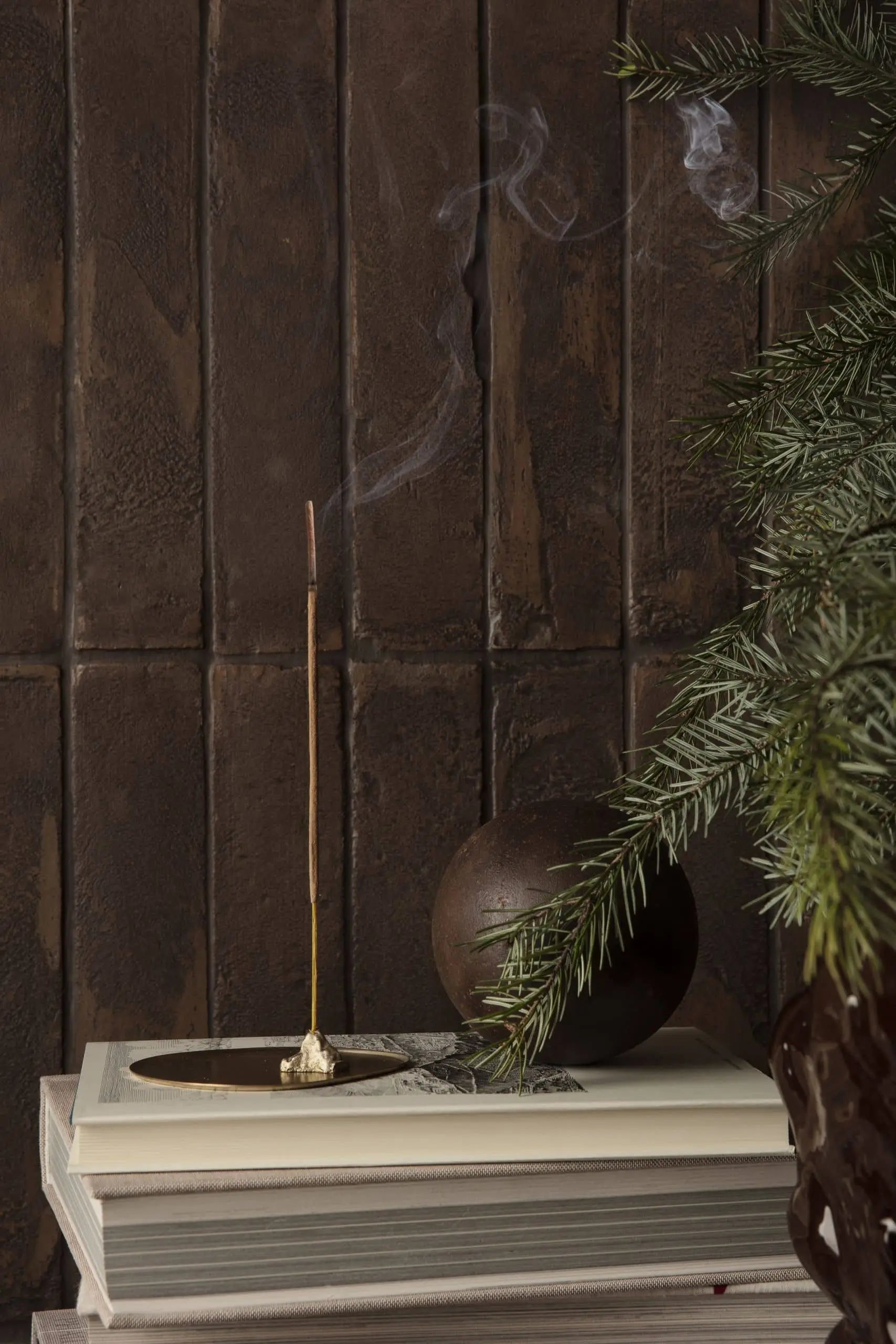 The ferm LIVING Stone Incense Burner by ferm LIVING holds a lit incense stick atop a closed book, styled with pine branches and a round brown accent against a dark wooden wall for peaceful, minimalist Zen decor.