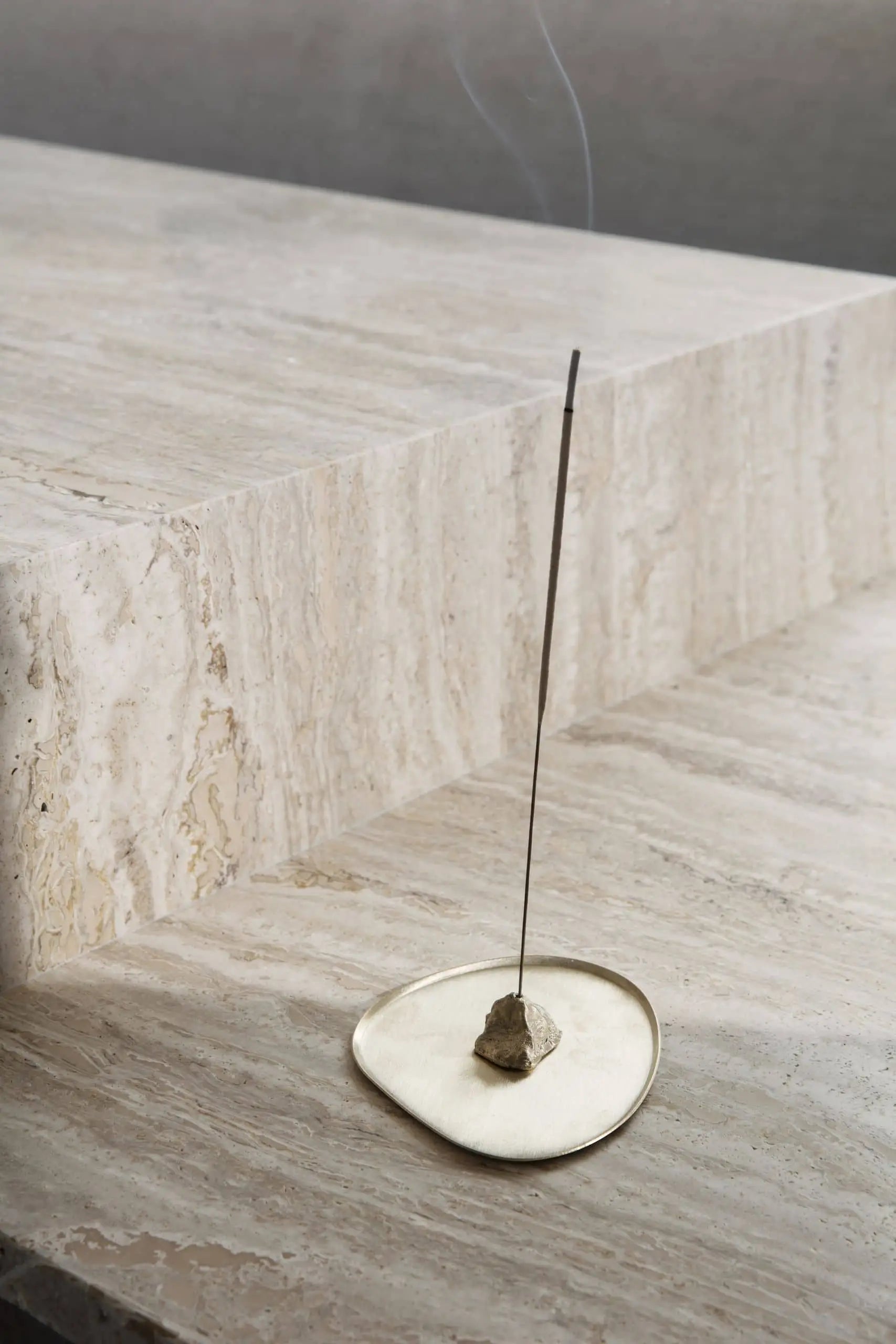 The ferm LIVING Stone Incense Burner holds an incense stick upright on a sleek, beige stone surface, with gentle smoke rising—an elegant piece by ferm LIVING to bring calm and style to your Zen-inspired decor.