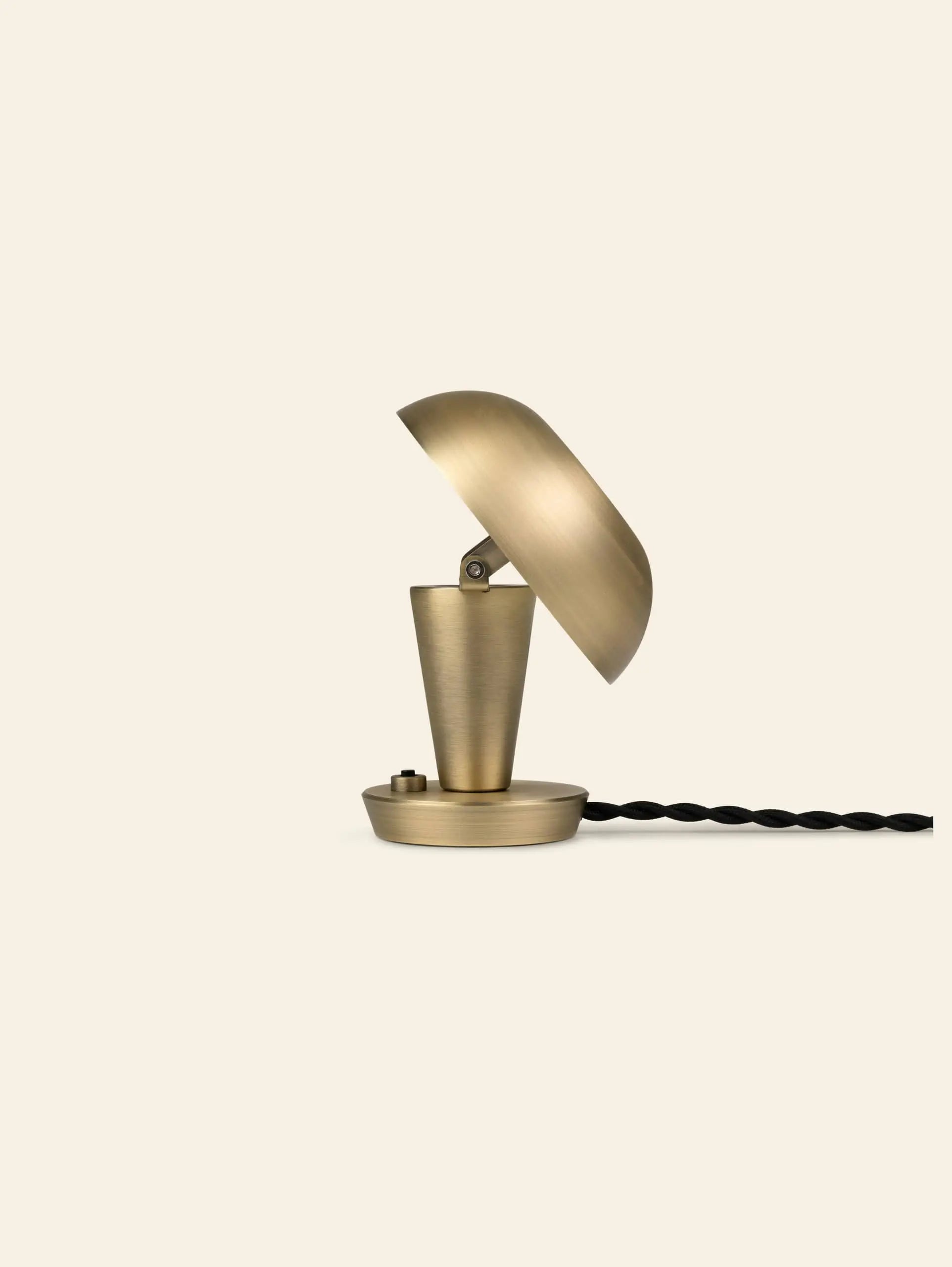 The ferm LIVING Tiny Lamp by ferm LIVING is a small, modern brass desk lamp featuring a dome-shaped shade, conical base, and black twisted cord, shown against a plain beige background.