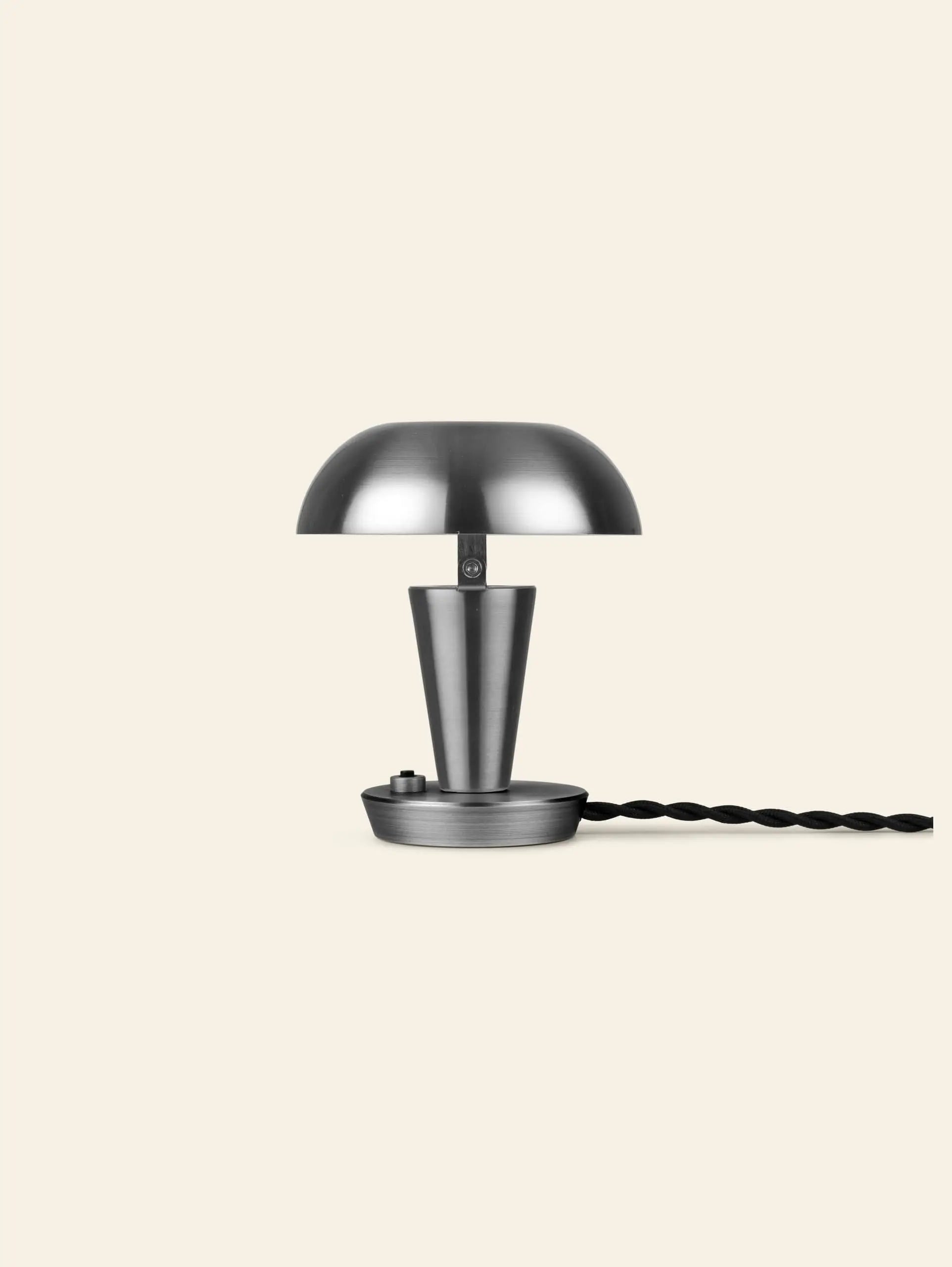 The ferm LIVING Tiny Lamp by ferm LIVING is a modern, minimalist metal table lamp featuring a dome-shaped shade, conical base, and twisted black power cord, set against a plain beige background.