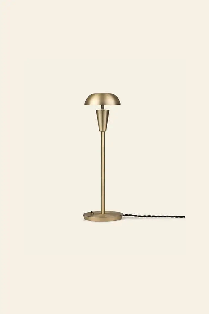 The ferm LIVING Tiny Table Lamp showcases modern sophistication with a brass dome shade, slender stem, round base, and black twisted cord—all set against a plain light beige background.