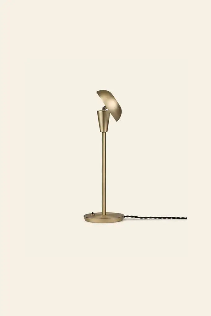 The ferm LIVING Tiny Table Lamp showcases modern sophistication with a brass finish, round base, slim stem, and angled dome-shaped shade. Its black power cord is visible against a plain beige background.