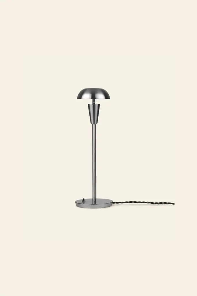 The ferm LIVING Tiny Table Lamp pairs modern sophistication with minimalist style, featuring a dome-shaped shade, slender stem, round base, and black twisted cord—all set against a plain beige background.