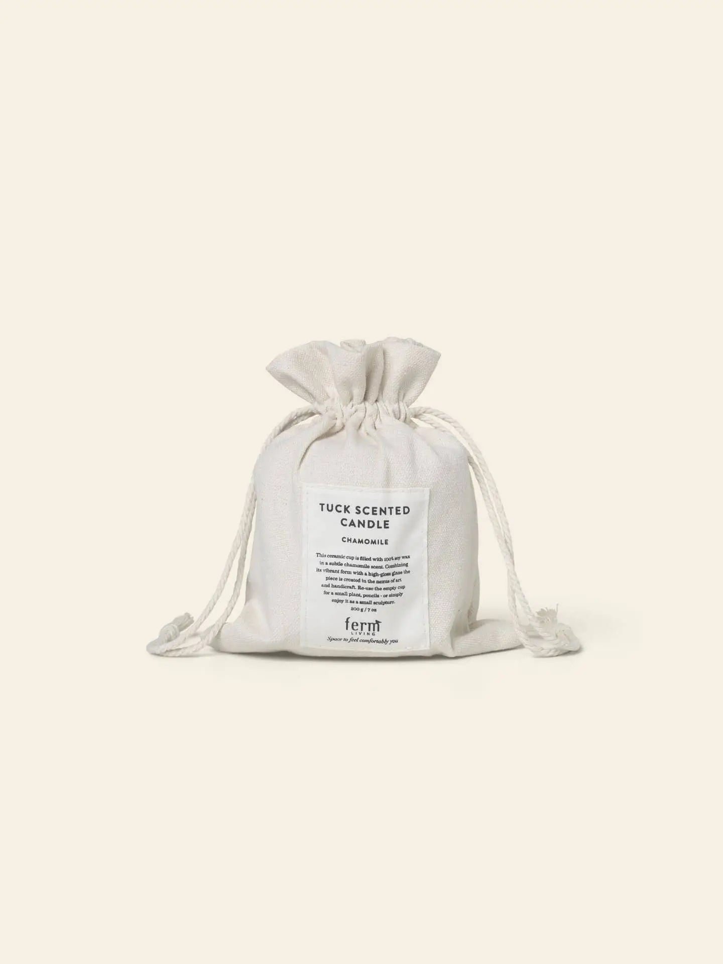 A small white drawstring pouch labeled “ferm LIVING Tuck Scented Candle,” featuring chamomile scent details for a calming ambiance, rests against a plain light background.