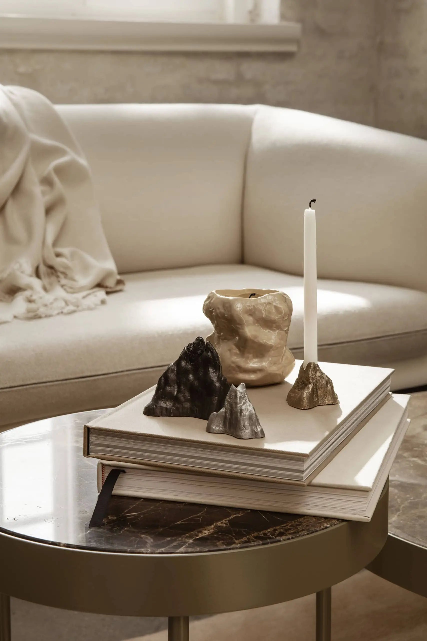 A modern living room features a light beige sofa with a draped blanket. On the round glass coffee table, stacked books, stoneware glazed sculptures, and the ferm LIVING Tuck Scented Candle create a calming ambiance.