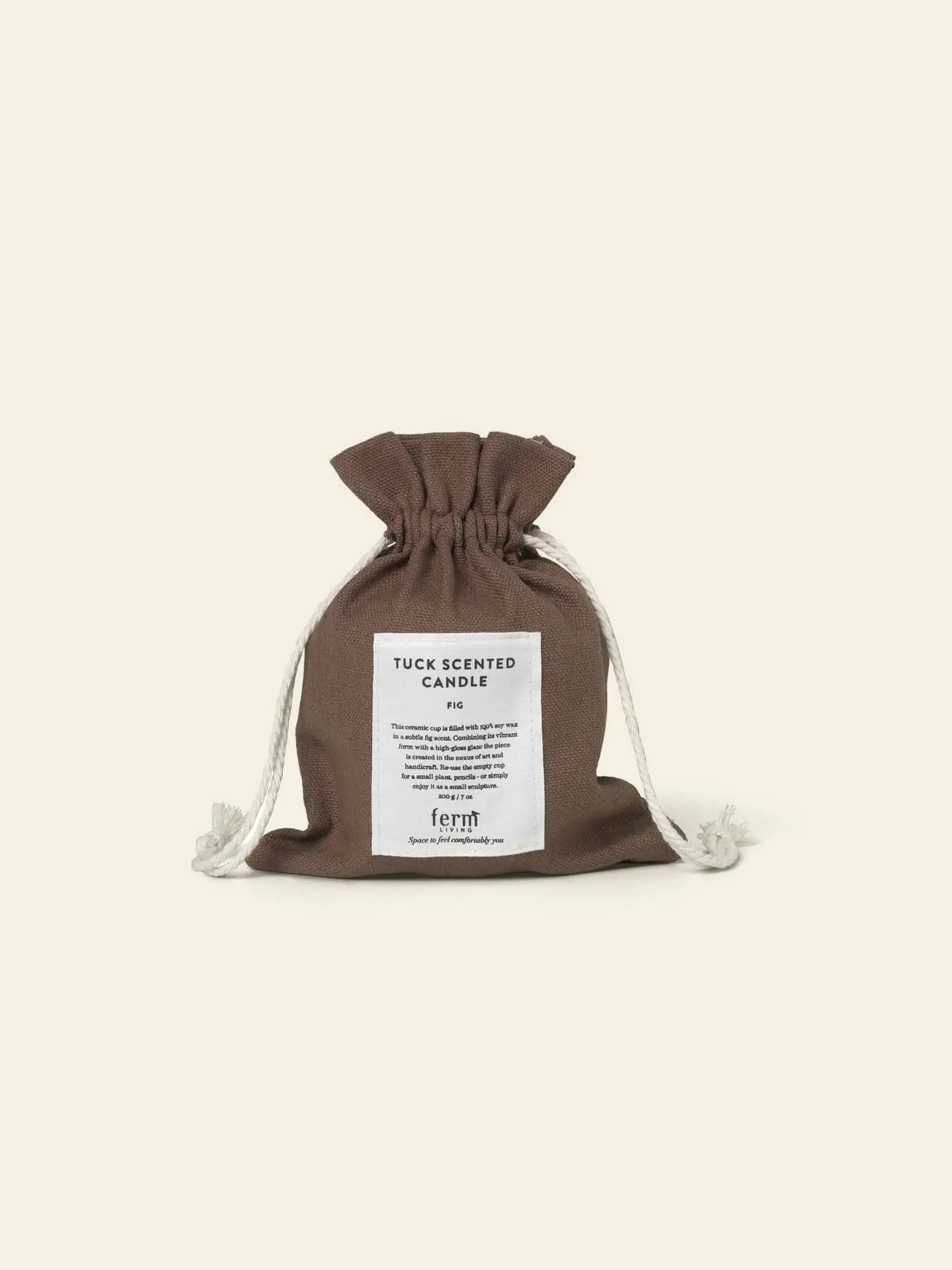 A brown drawstring pouch with white cords sits upright. A white front label reads “ferm LIVING Tuck Scented Candle,” evoking a calming ambiance. The background is a soft beige.