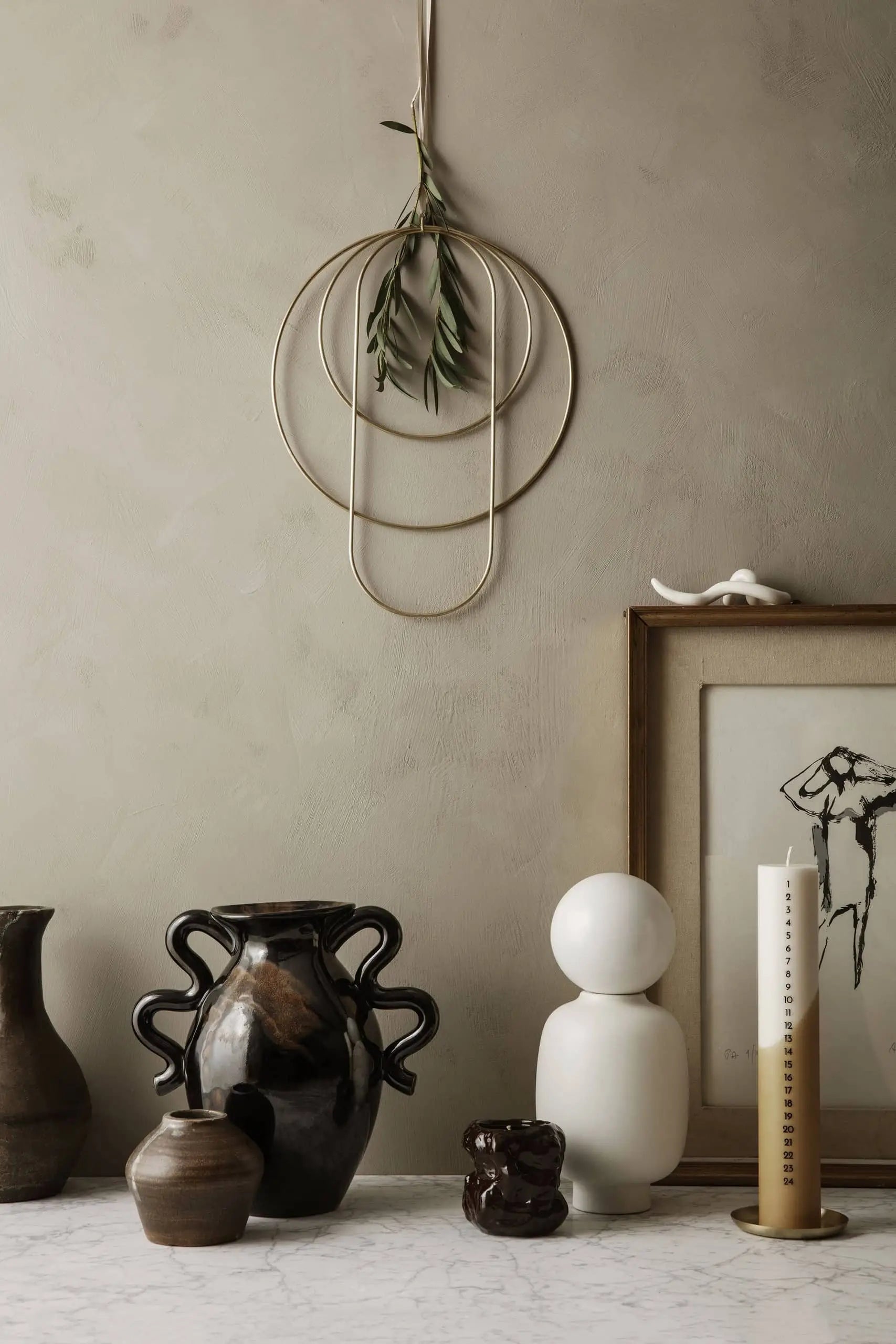 A minimalist decor featuring ceramic vases, abstract sculptures, a framed sketch, greenery with a gold wall hanging, and the ferm LIVING Tuck Scented Candle on marble, all set against a neutral-toned wall for a calming ambiance.