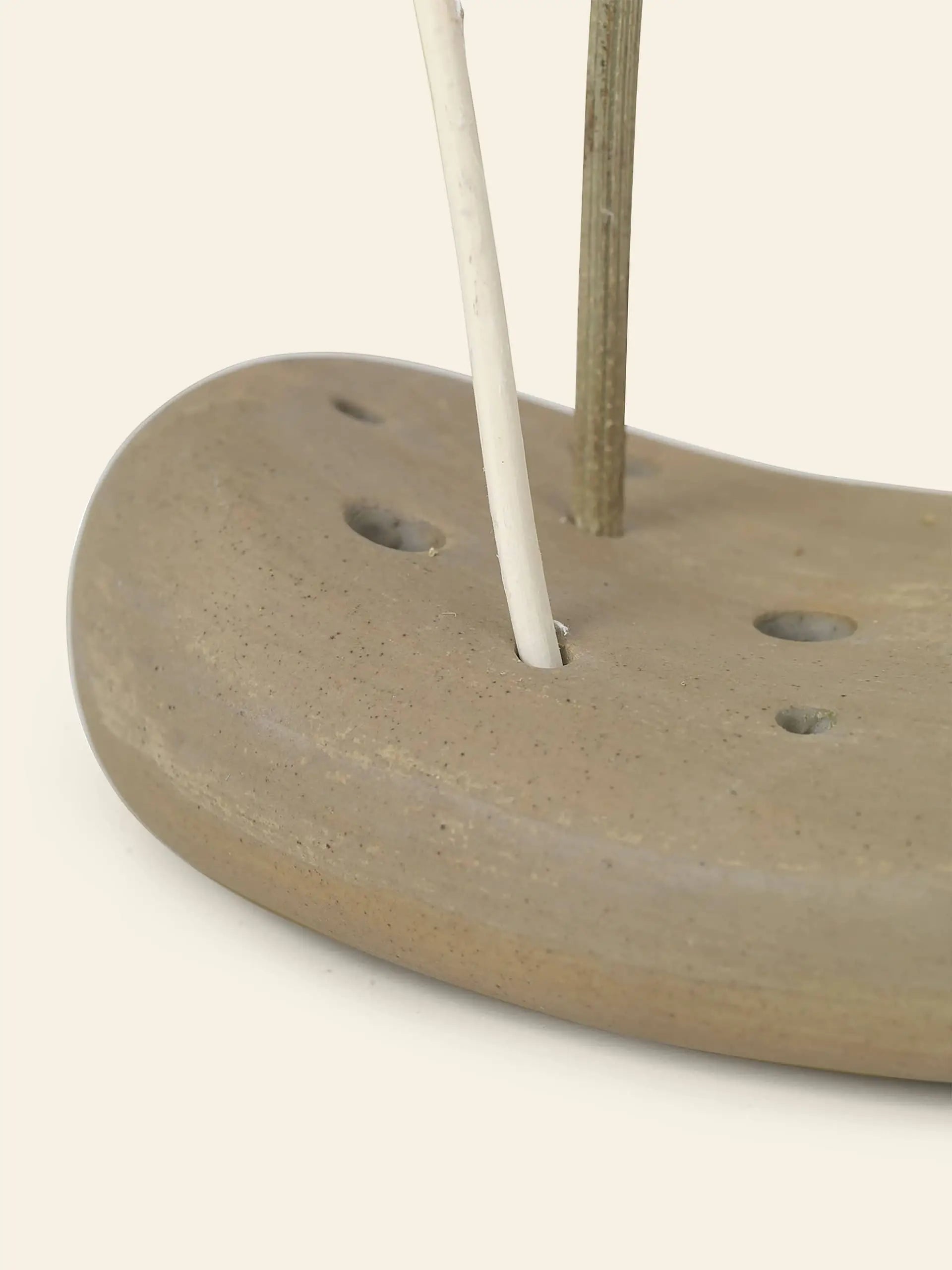 A close-up of the ferm LIVING Vanitas Flower Stone by ferm LIVING, an elegant glazed porcelain holder with multiple holes, displaying two slim sticks—one white and one brown—on a plain background. Ideal for modern decor.