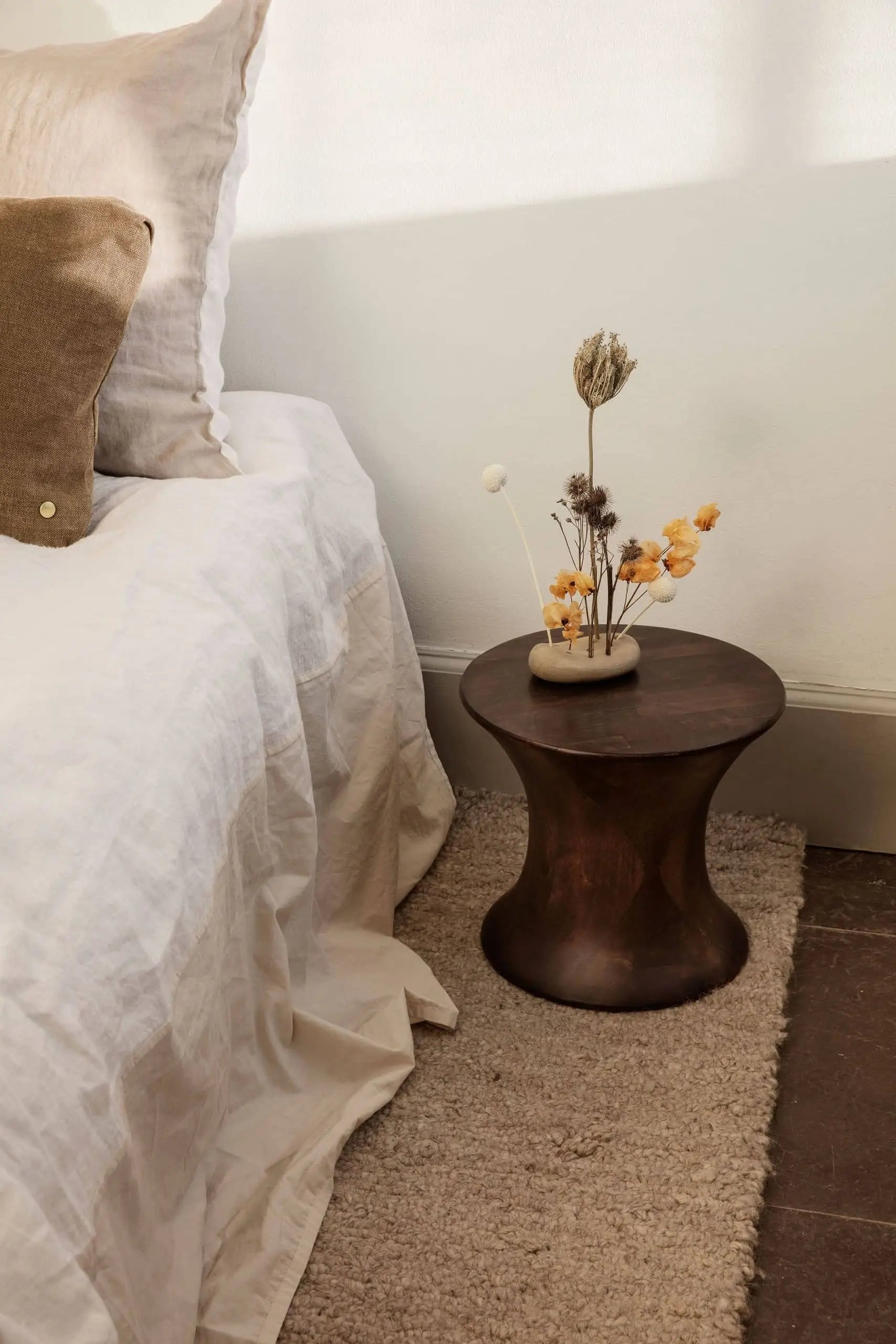 A cozy bedroom scene with a white bedspread, brown pillow, and dark wooden side table featuring the ferm LIVING Vanitas Flower Stone vase with dried flowers, all on a beige textured rug.