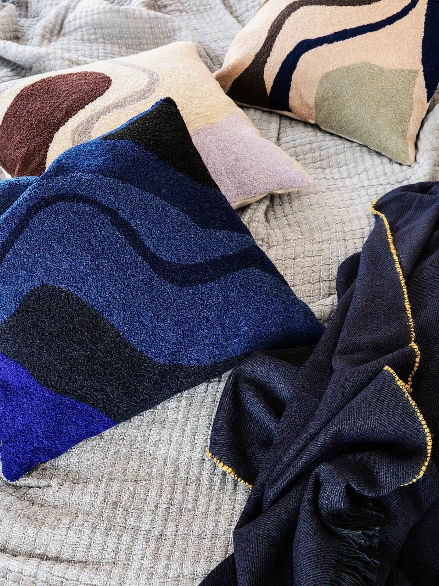 Three decorative pillows, including the ferm LIVING Vista Cushion featuring wave patterns in blue, beige, and brown, rest on a quilted gray blanket beside a dark blue throw with gold trim—ideal for a luxurious modern home.