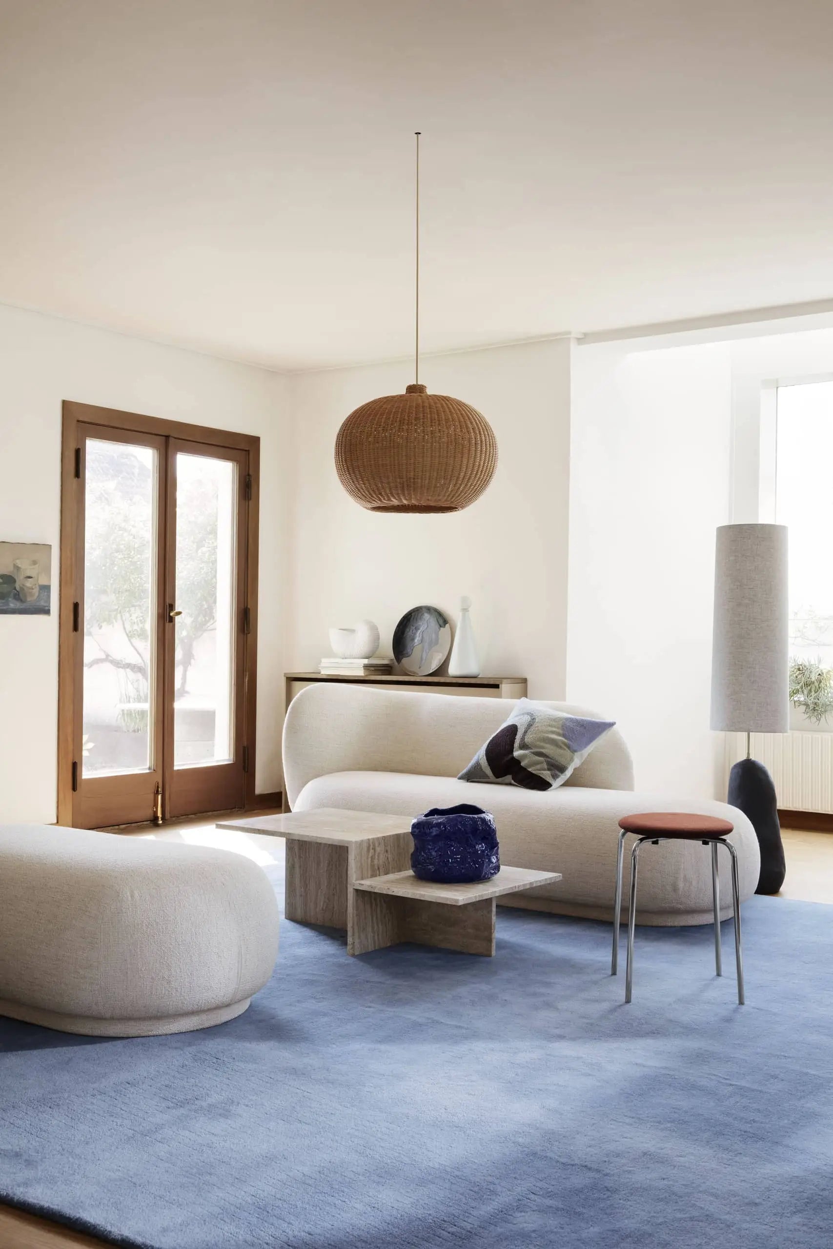 A modern luxury living room with cream, rounded furniture, the ferm LIVING Vista Cushion, a light blue rug, wooden coffee table with blue vase, tall floor lamp, woven pendant light, and natural light streaming through glass doors.