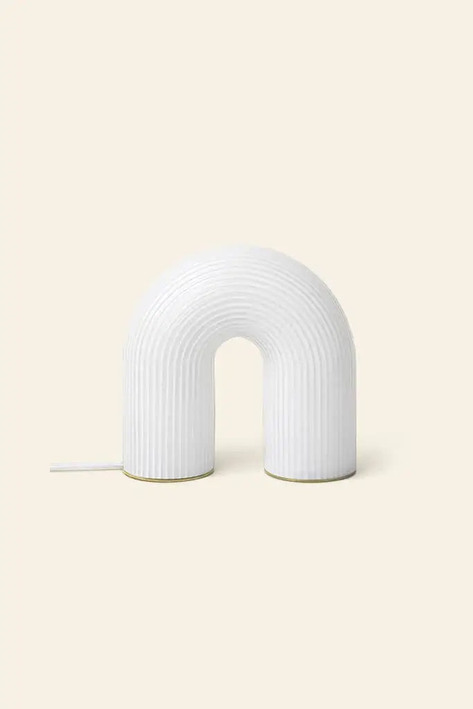The ferm LIVING Vuelta Lamp, a modern white U-shaped sculptural lamp with a ribbed surface, sits on a light beige background. A cord extends from the left, emphasizing its elegant design inspired by ferm LIVING aesthetics.