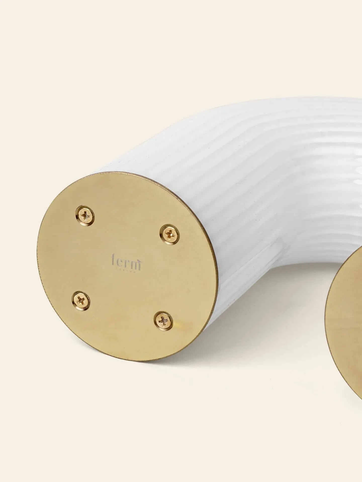 A close-up of the ferm LIVING Vuelta Lamp shows its sculptural, rippled design and a round gold metal end plate with ferm engraved, secured by four screws. The background is light cream. Brand: ferm LIVING.