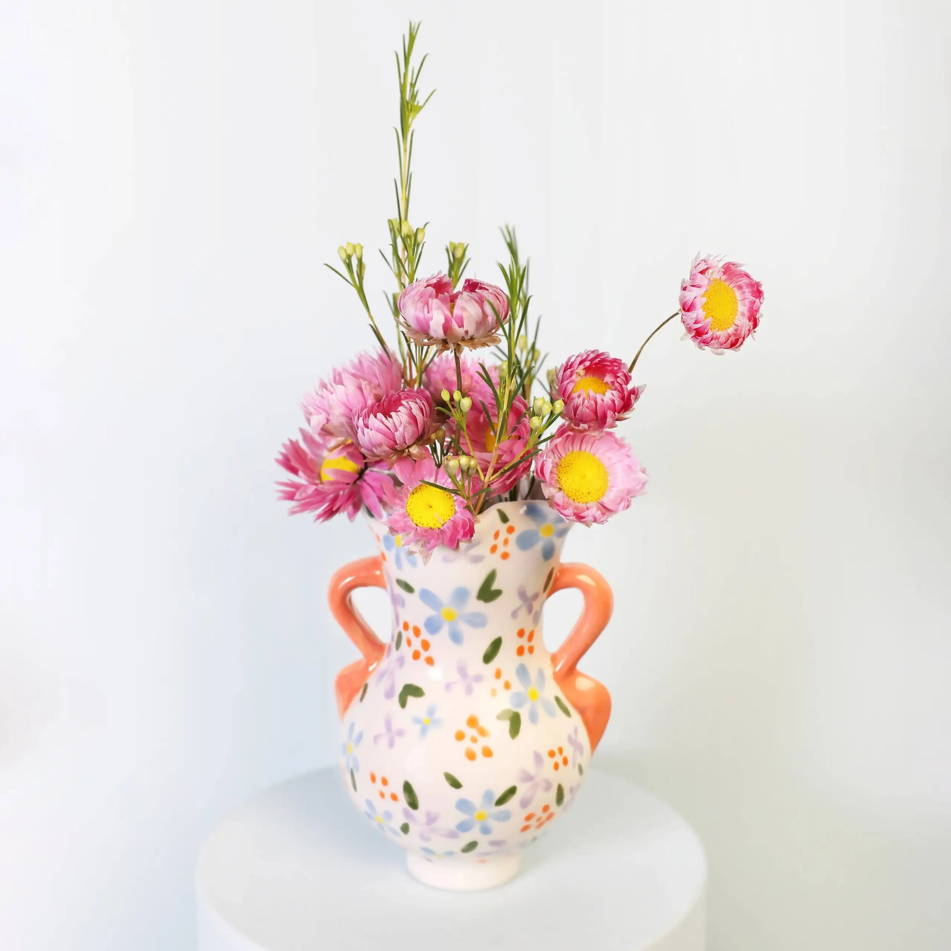 The &klevering Bloom Vase by &klevering features orange handles and multicolored floral patterns, displaying pink and yellow blooms for a romantic accent against a simple light backdrop.