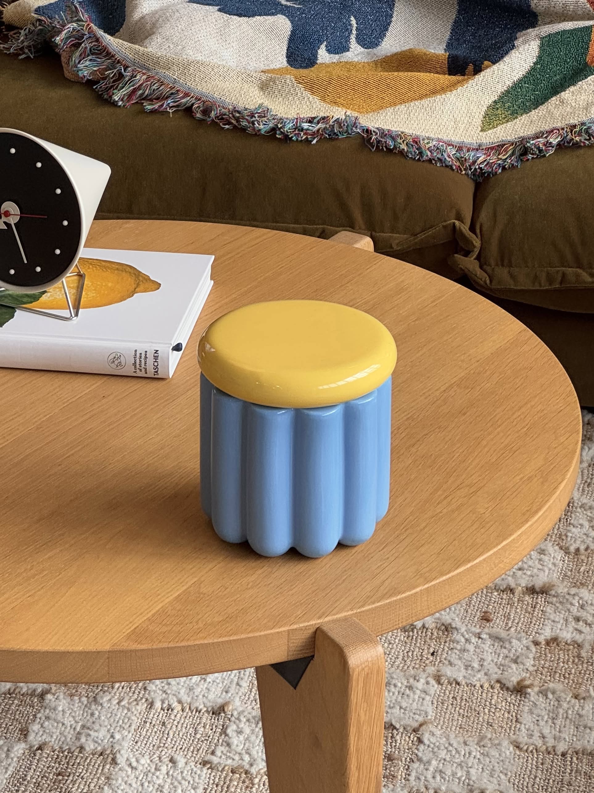 The &klevering Charlo Jar - Blue, featuring a blue body and yellow lid with an airtight silicone seal, adds a pop of color on the coffee table, with a book, modern clock, brown couch, and colorful blanket in the background.