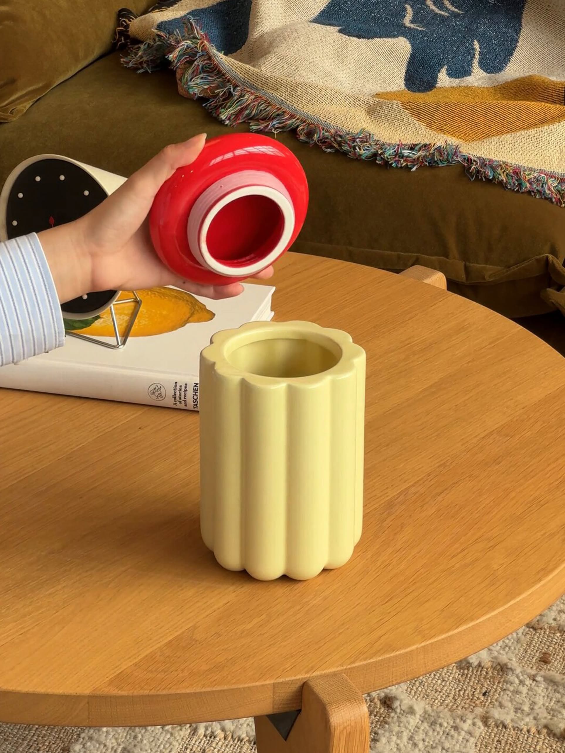 A hand holds the &klevering Charlo Jar - Off-White above a yellow fluted vase on a wooden table. A book and clock sit nearby, with a colorful blanket in the background—ideal for keeping treats fresh in style.