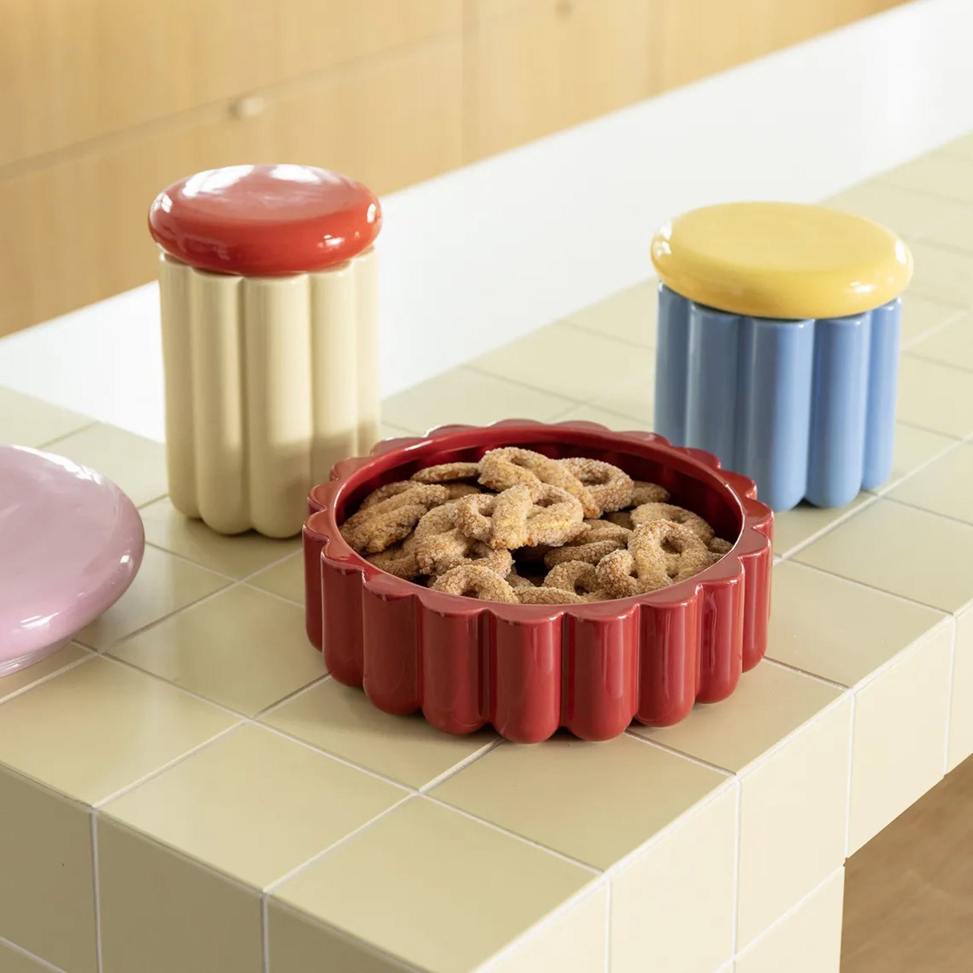 The &klevering Charlo Jar in off-white ceramic, featuring playful fluted design and a red lid, rests on tiled surfaces—perfect for storing sugar-coated cookies that delight like Charlotte cakes.