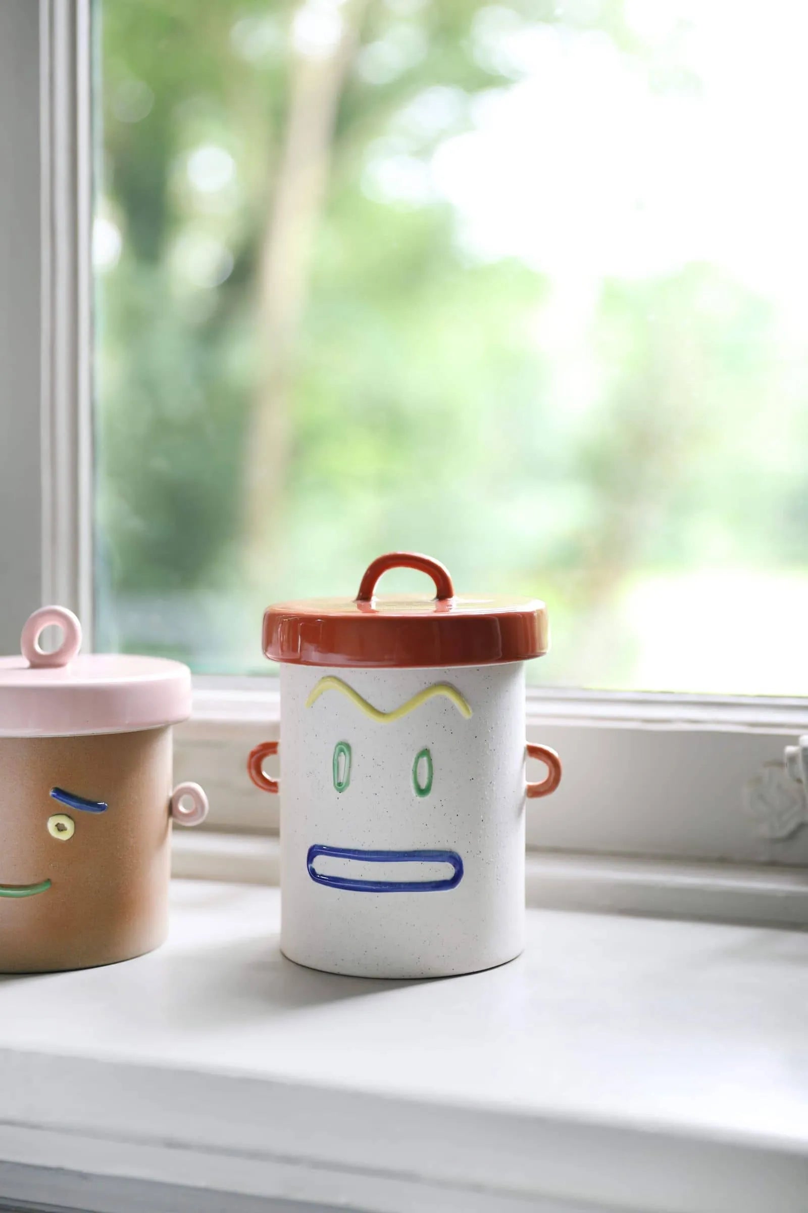 Add playful charm to your windowsill with &klevering Cheeky Jars—two decorative containers featuring cartoon faces, one beige with a pink lid and one white with a red lid—perfect storage from &klevering against a lush, green backdrop.