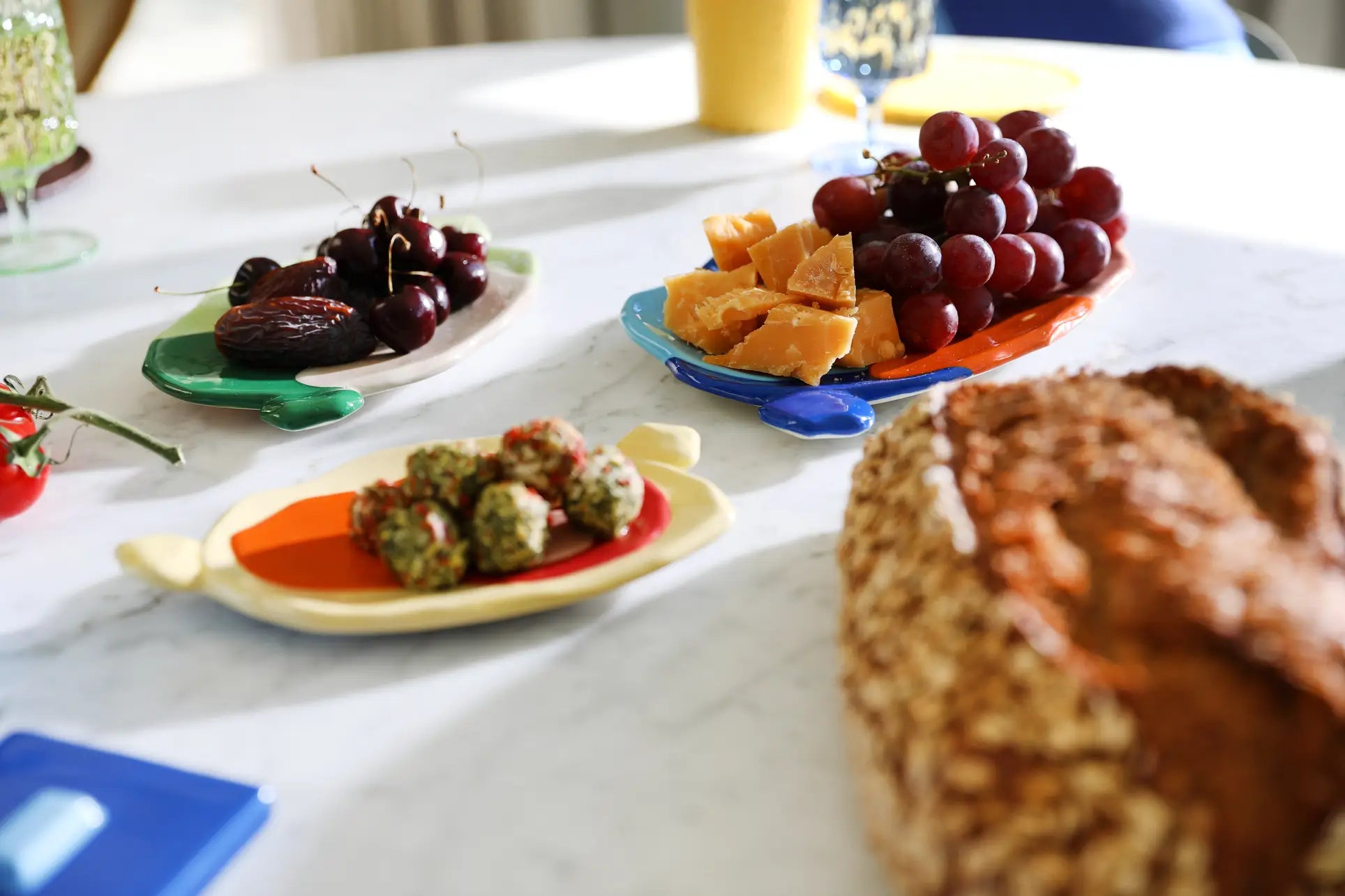 Displayed on a white table, cherries, dates, grapes, cheese, bread, and appetizers are artfully arranged on the &klevering Chunky Platter, Medium by &klevering—its bold colors and fish shape add artistic charm in natural light.