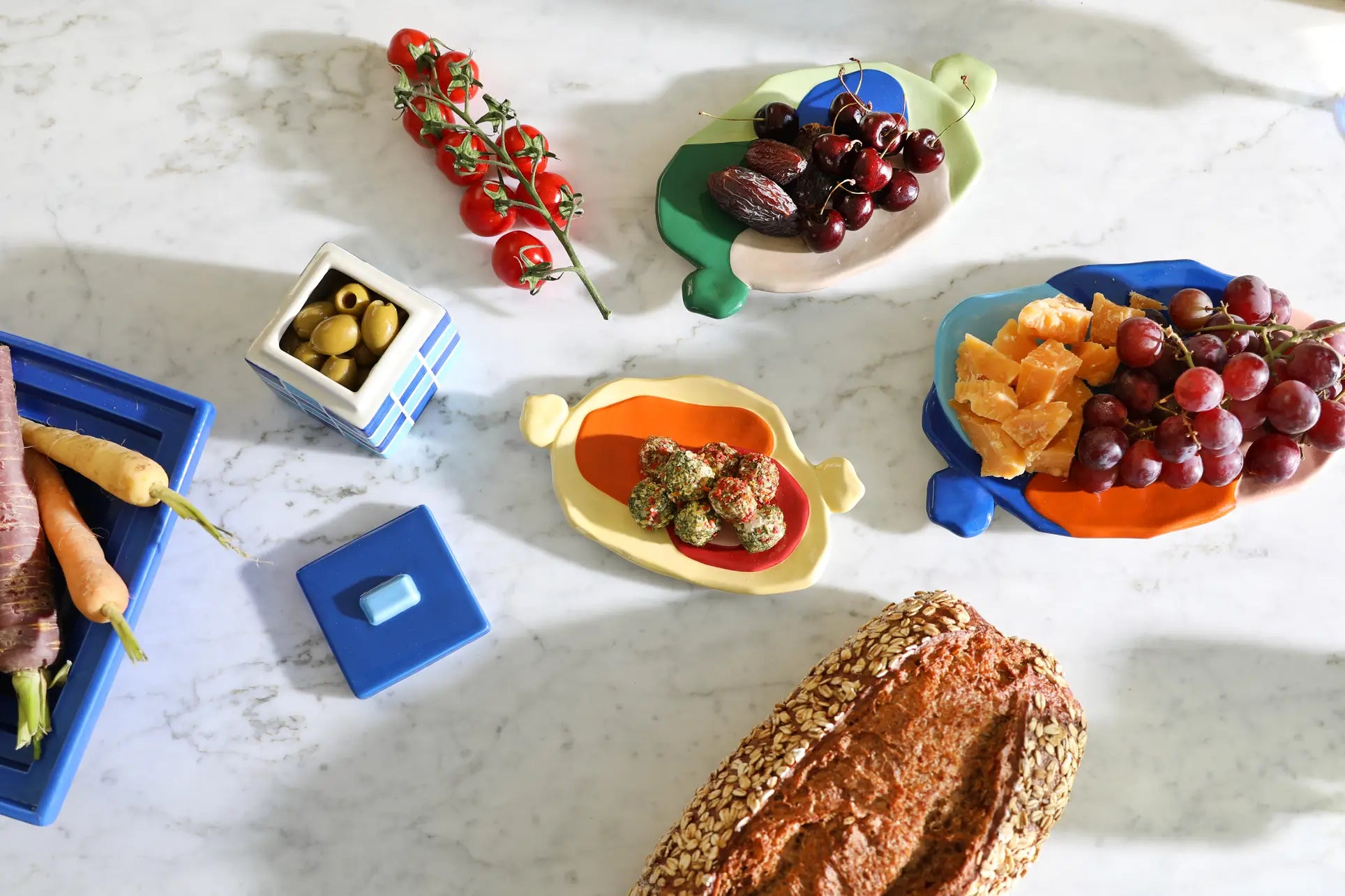 A marble surface displays fresh fruits, veggies, cheese, bread, vine tomatoes, green olives, and a seeded loaf on the vibrant &klevering Chunky Platter (Medium), adding artistic charm and bold color to the scene.
