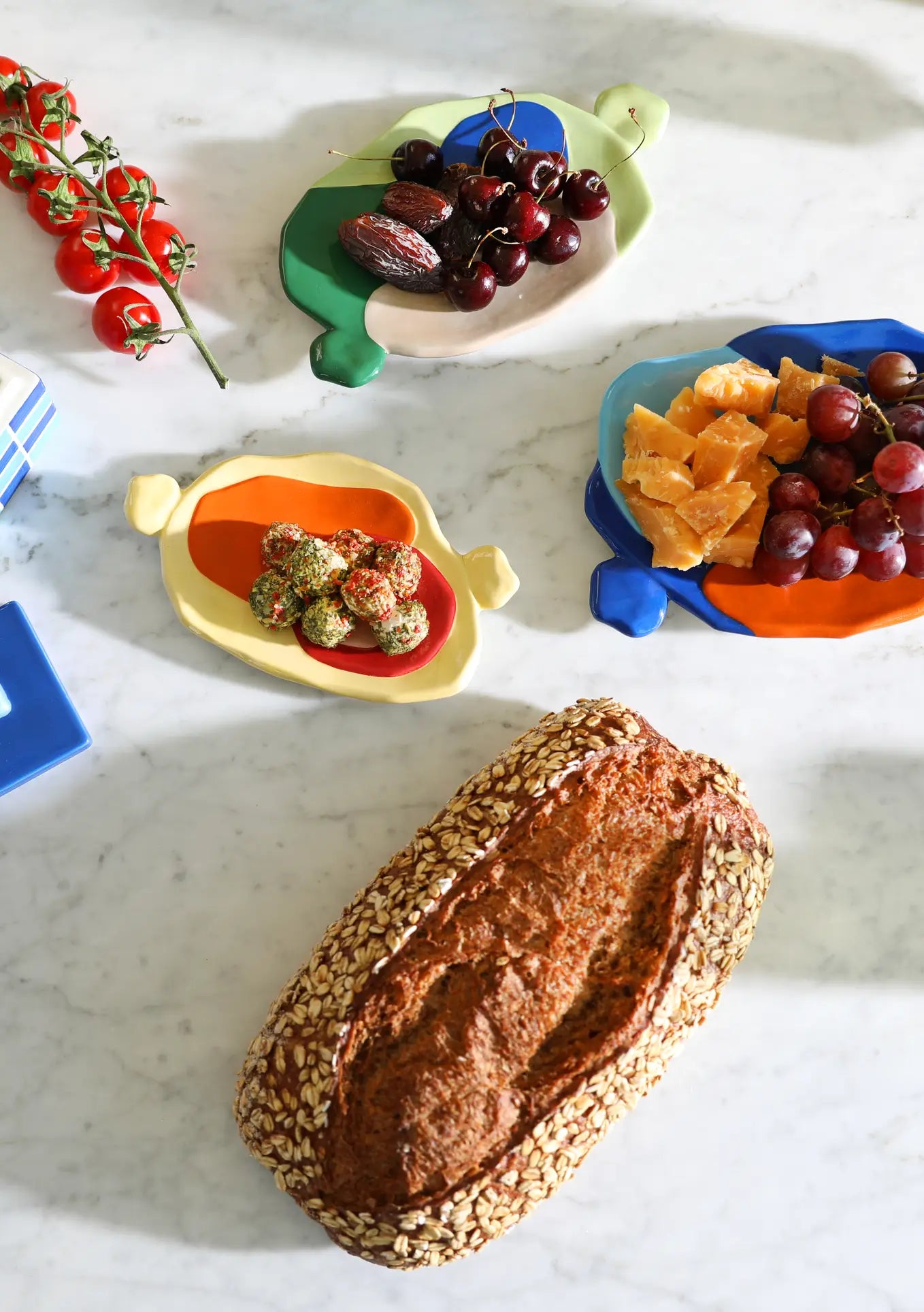 A loaf of seeded bread, red grapes, orange slices, dates, cherries, vine tomatoes, and cheese balls are arranged on a marble surface with the &klevering Chunky Platter, Small—showcasing bold colors and artistic charm.