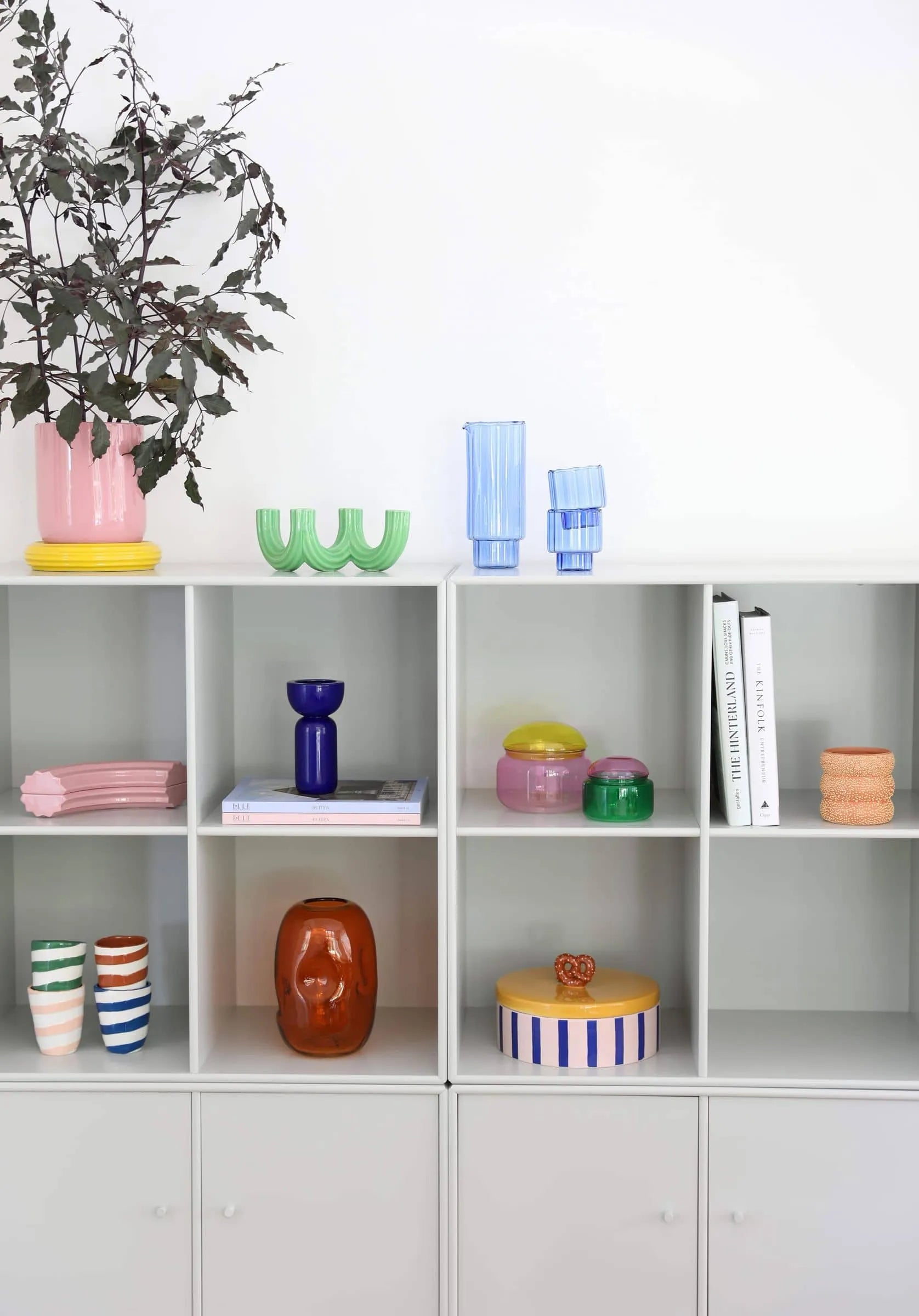 A white shelving unit displays colorful vases, bowls, unique decor accents, books, and the &klevering Churros Candle Holder Green by &klevering, plus a pink vase with leafy branches on top, all set against a plain white wall.