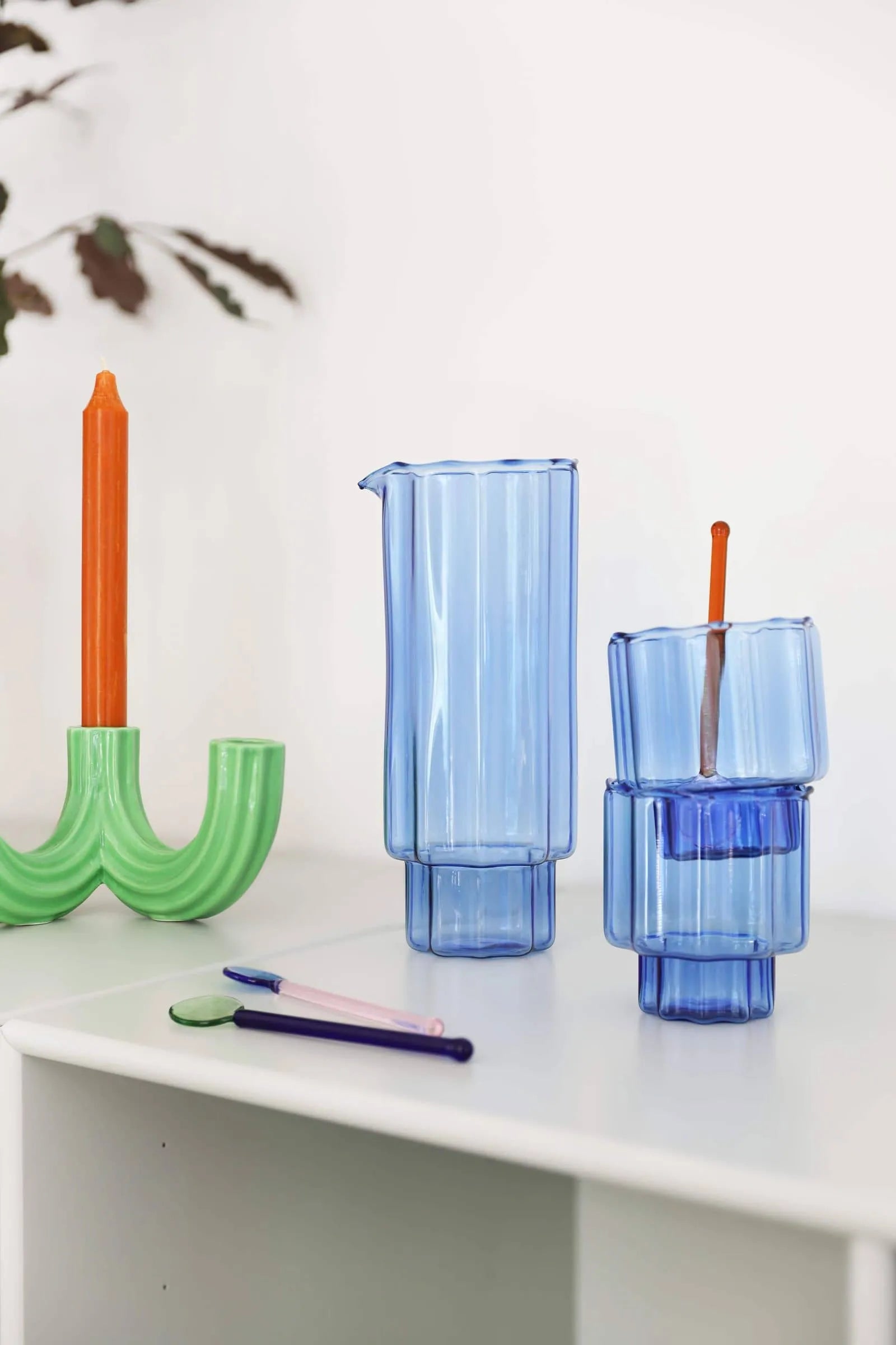 A blue glass pitcher and two stacked blue glasses sit on a white surface next to an orange candle in the &klevering Churros Candle Holder Green, two pens, and a leafy branch for a charming home decor scene.