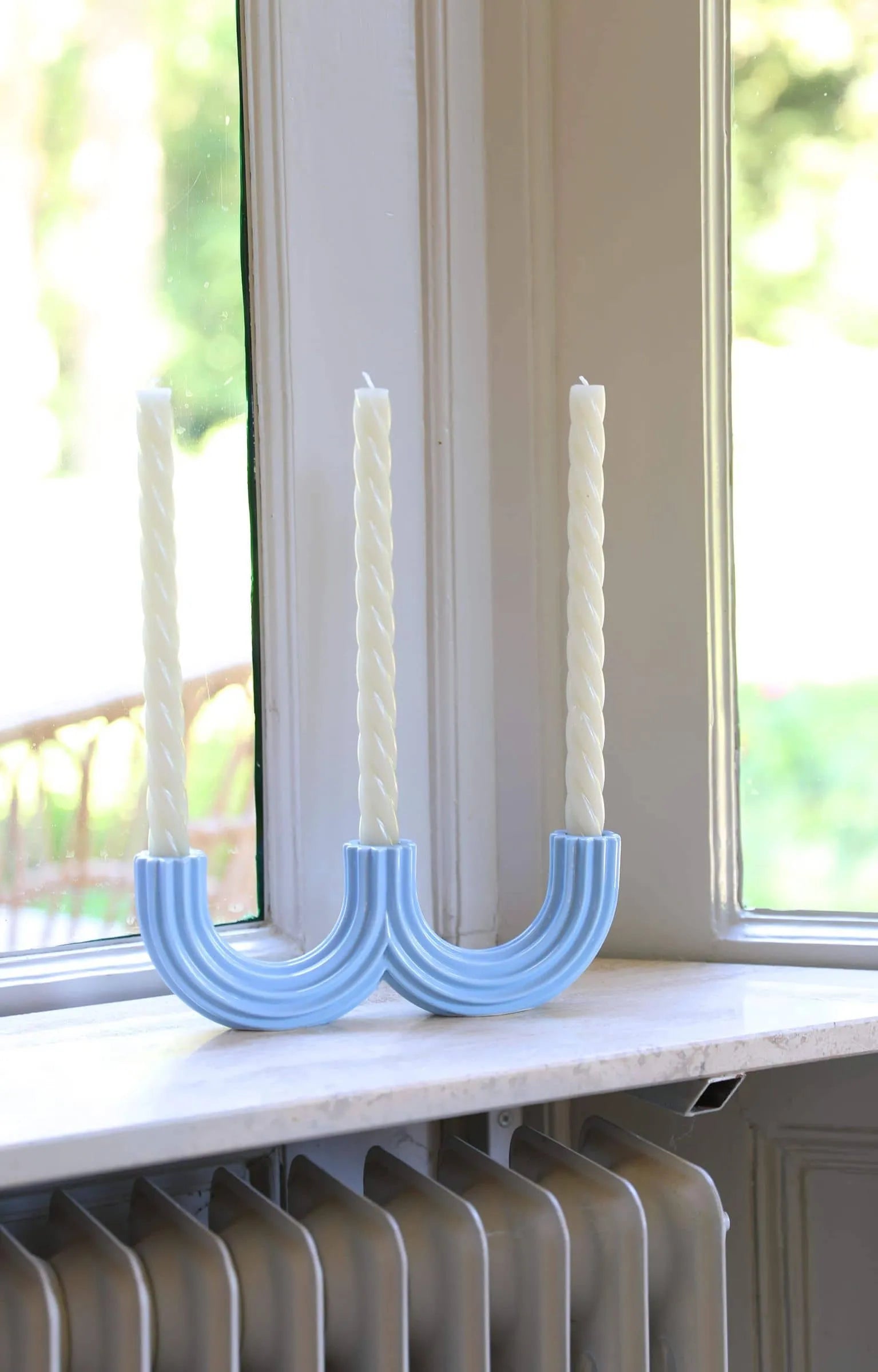 Two white twisted candles rest in &klevering Churros Candle Holder Light Blue by &klevering on a windowsill. Sunlight streams in, highlighting the handcrafted holders and lush greenery outside—an elegant home decor accent for any space.
