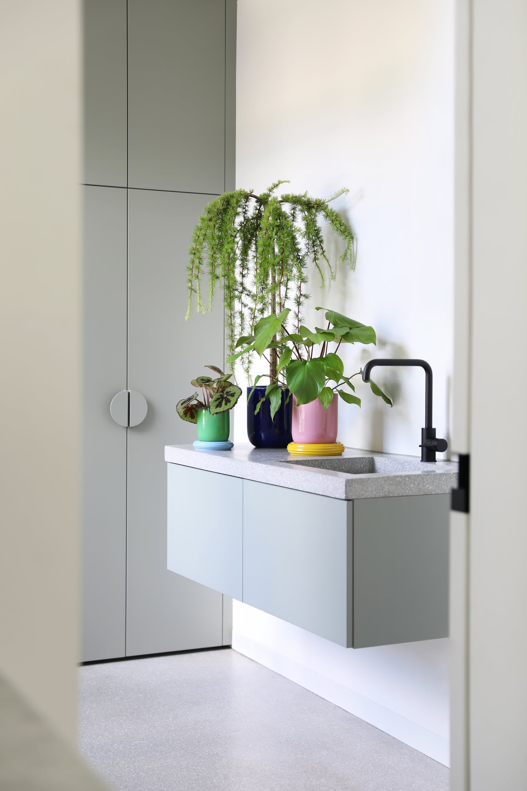 A modern, minimalist bathroom features a floating gray vanity with a black faucet, topped with the cheerful &klevering Churros Planter for a pop of color against white walls and tall gray cabinets.