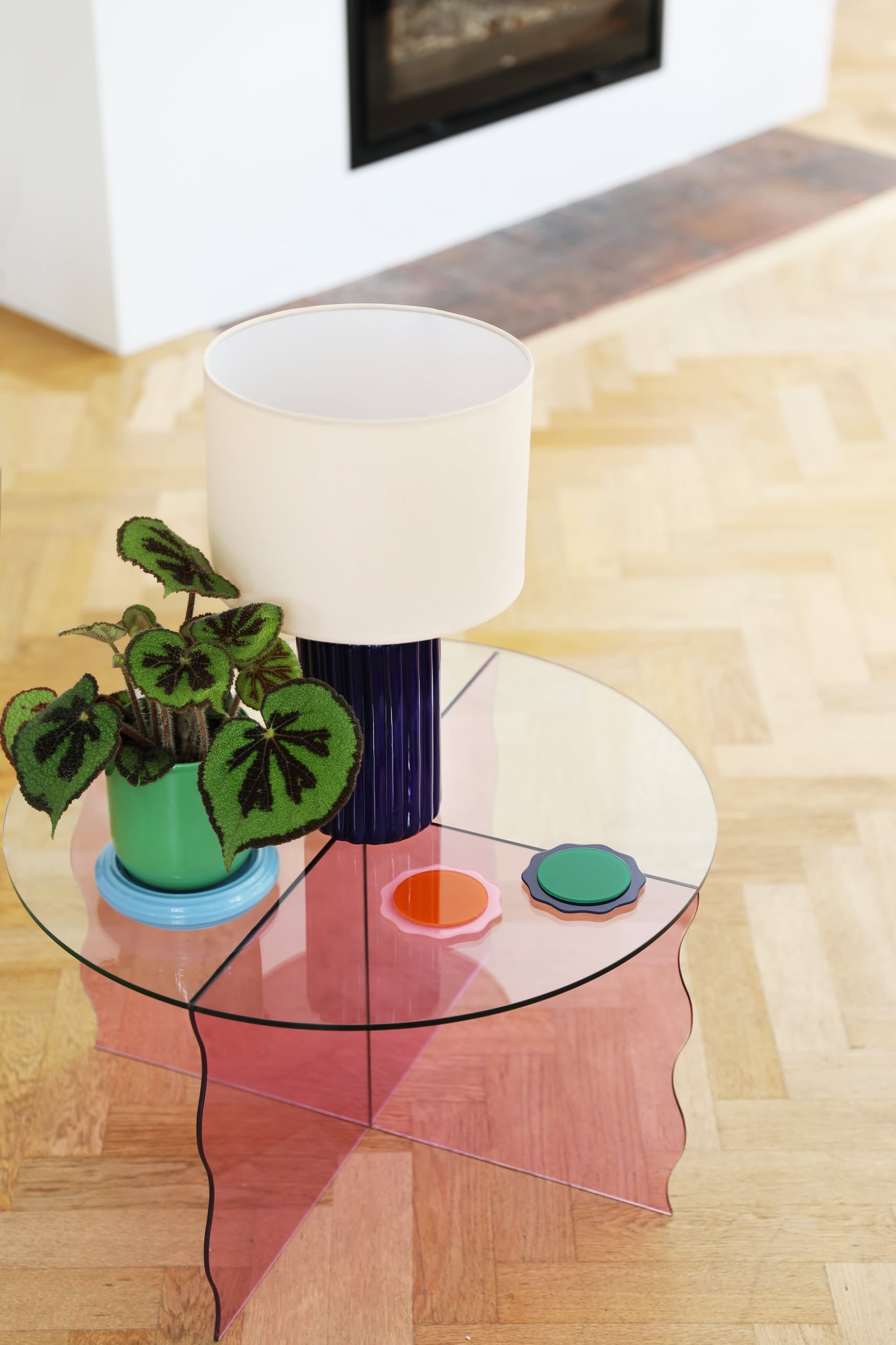 A round glass coffee table with a pink base holds a white lamp with a blue base, an &klevering Churros Planter by &klevering, and two colorful coasters, all set on light wood parquet flooring near a fireplace.