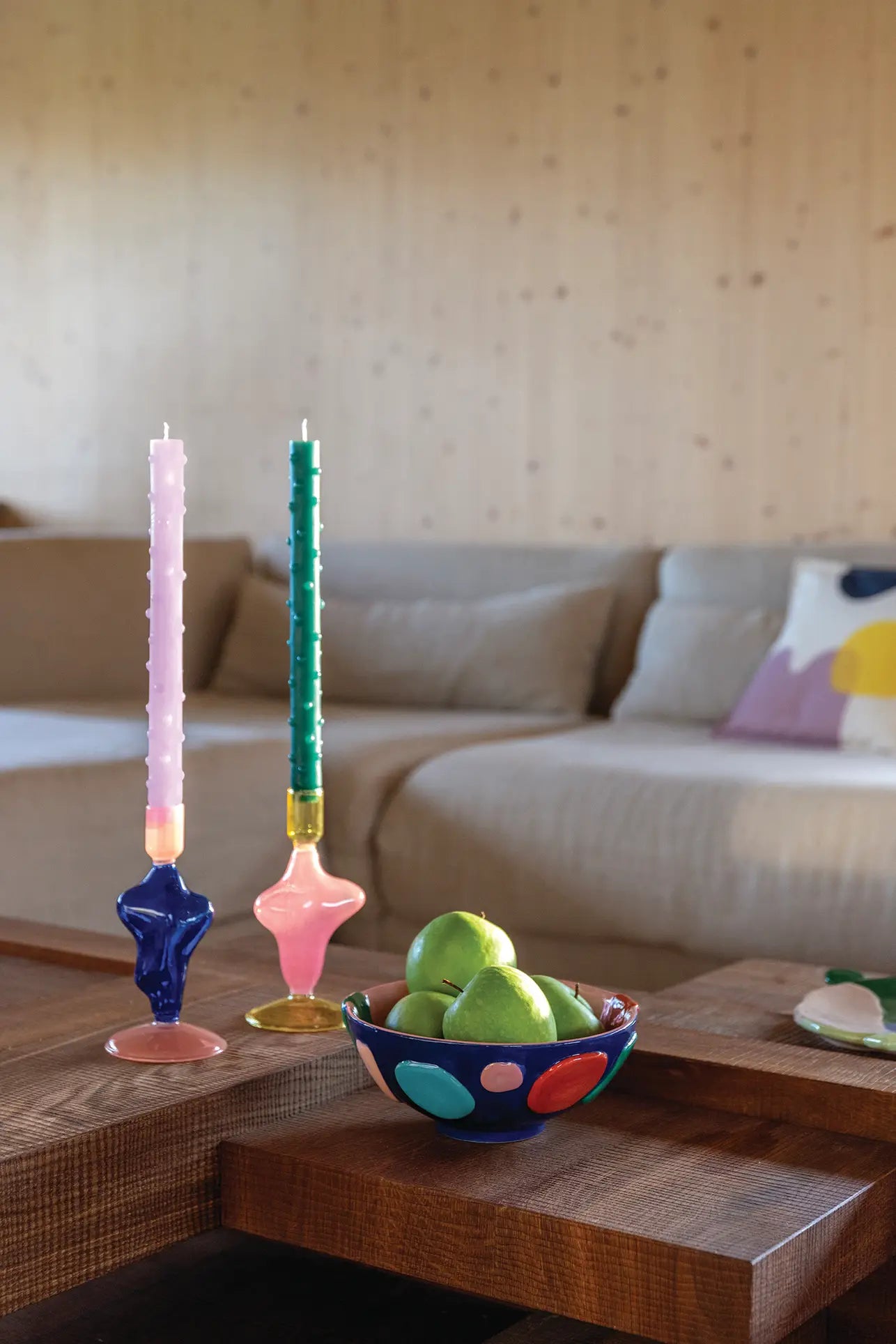 A wooden coffee table displays the &klevering Crafty Bowl, Small filled with green apples and two tall, colorful candle holders. A beige sofa with a patterned pillow sits behind, adding comfort and modern style to the room.