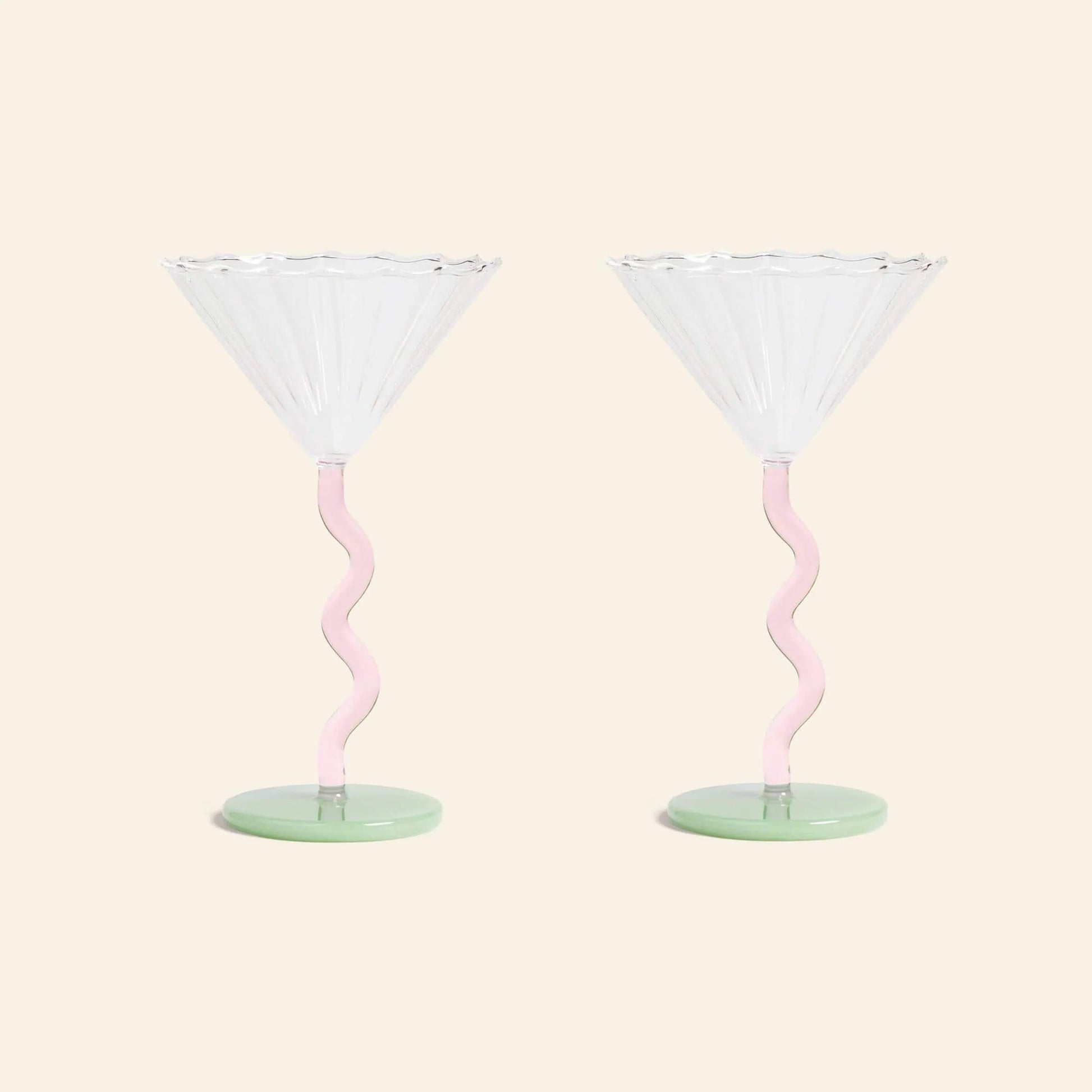 The &klevering Curved Coupe Glass Set of 2 features clear, scalloped rims, pink wavy stems, and green bases—charming colored glassware by &klevering, ideal for celebratory gatherings against a plain beige backdrop.