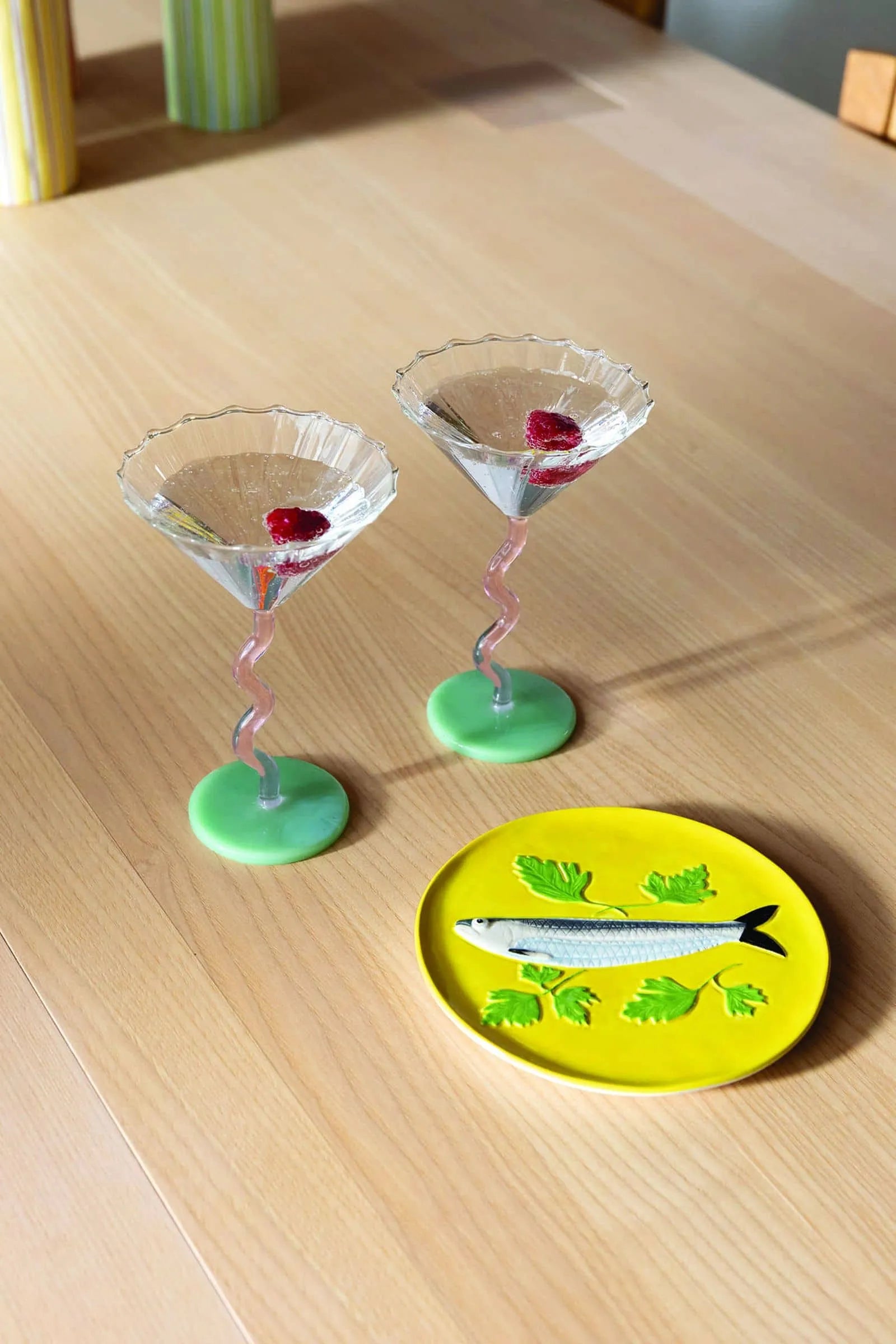 The &klevering Curved Coupe Glass Set of 2, featuring pink twisted stems and green bases, holds drinks with red garnish on a light wooden table beside a yellow fish-decorated plate with green leaves. Perfect for celebrations.