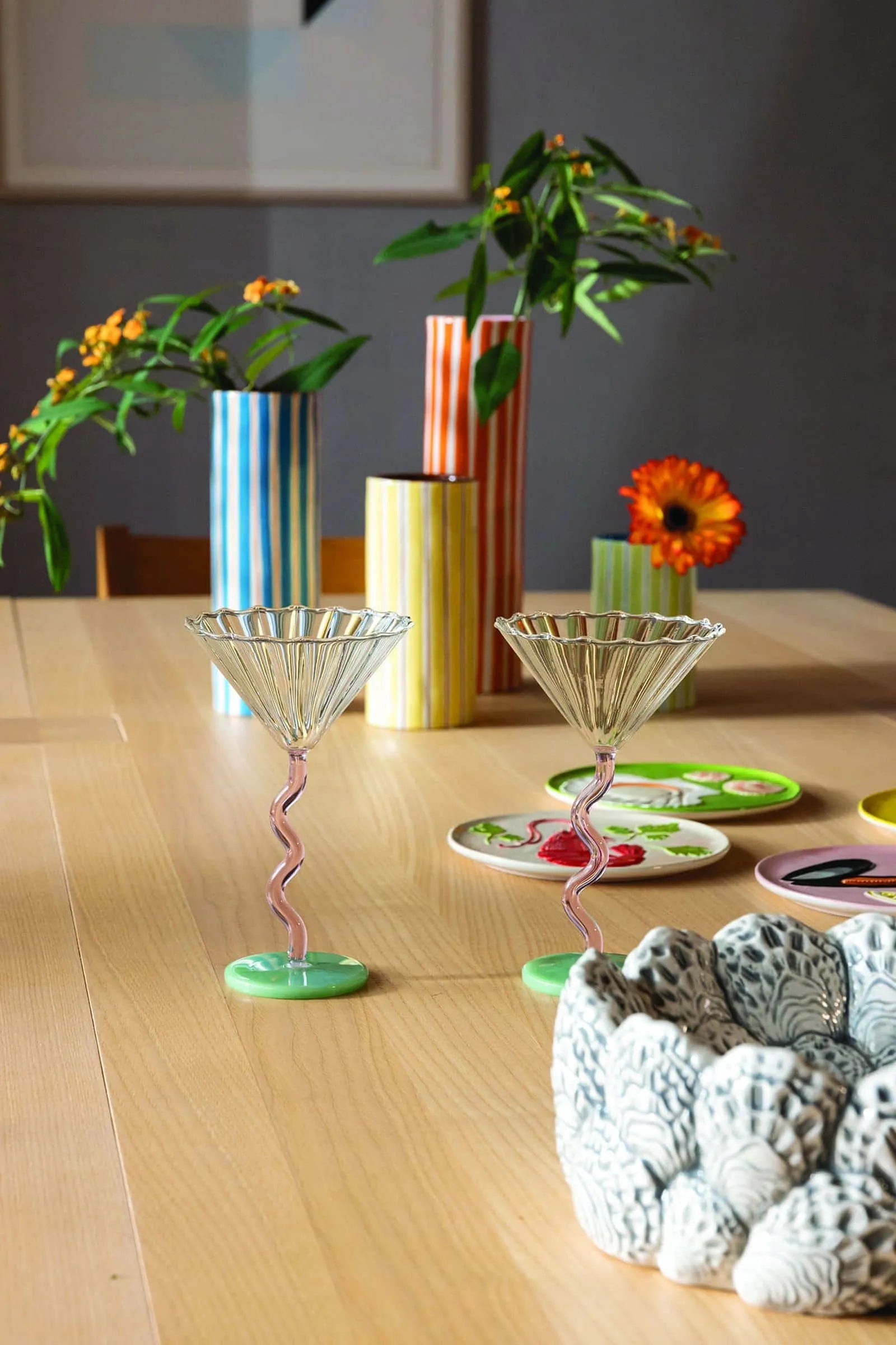 Two &klevering Curved Coupe Glasses (set of 2) from &klevering, with playful twisted stems, rest on a wooden table amid striped vases, floral accents, and colorful plates—perfect for lively gatherings. A textured patterned bowl appears in the foreground.