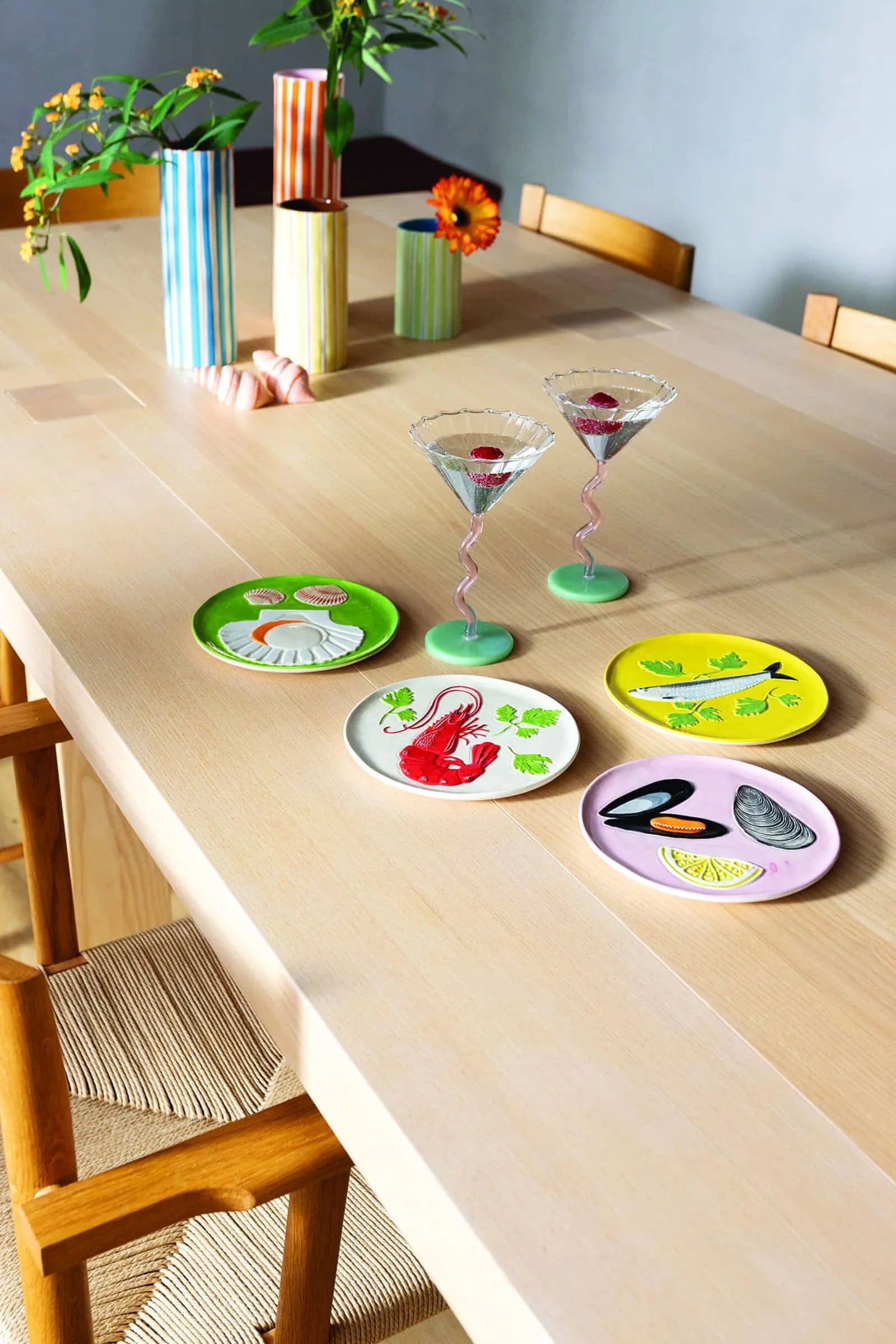 A light wooden dining table set for a celebration with four seafood-themed plates and two &klevering Curved Coupe Glasses, surrounded by wooden chairs and striped vases filled with flowers in the background.