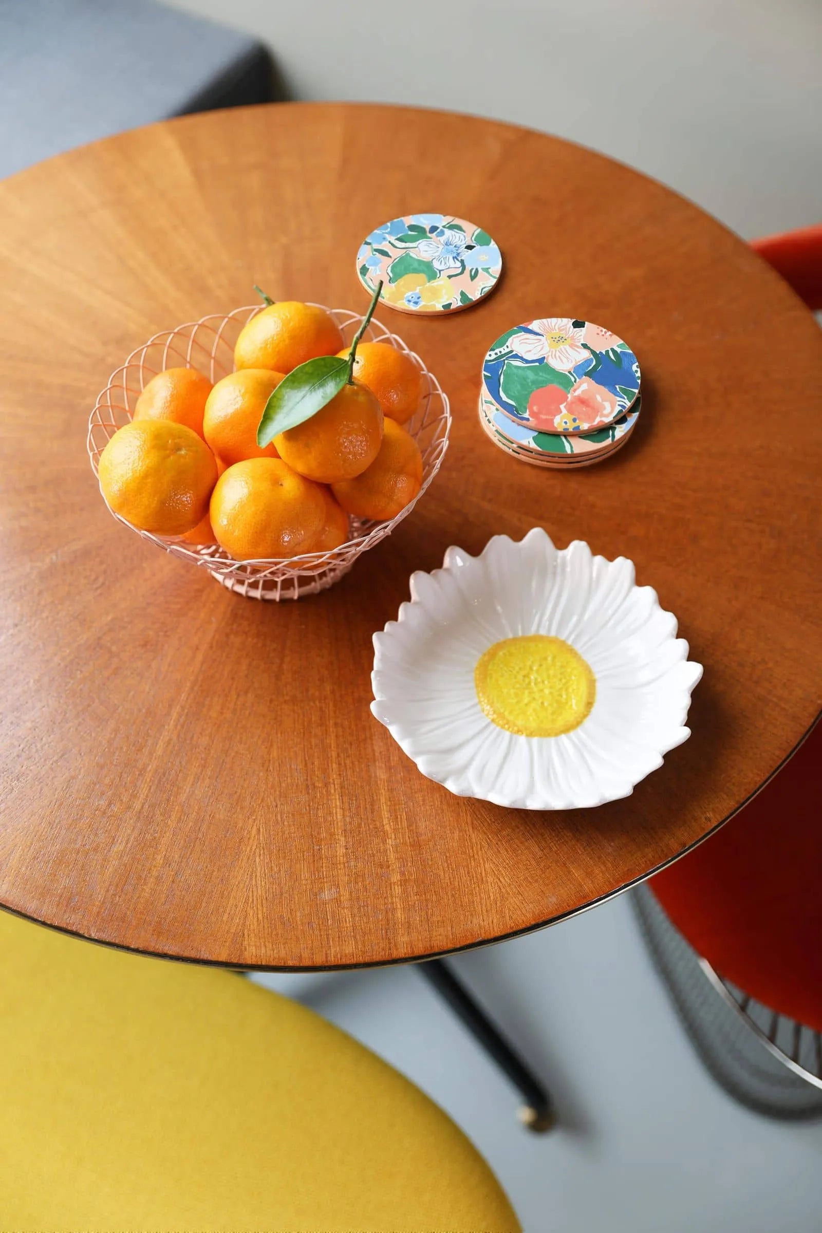 A round wooden table set for a garden dinner features the &klevering Daisy Bowl by &klevering filled with oranges, a daisy-shaped plate, three colorful coasters, and yellow, red, and gray chairs surrounding the scene.