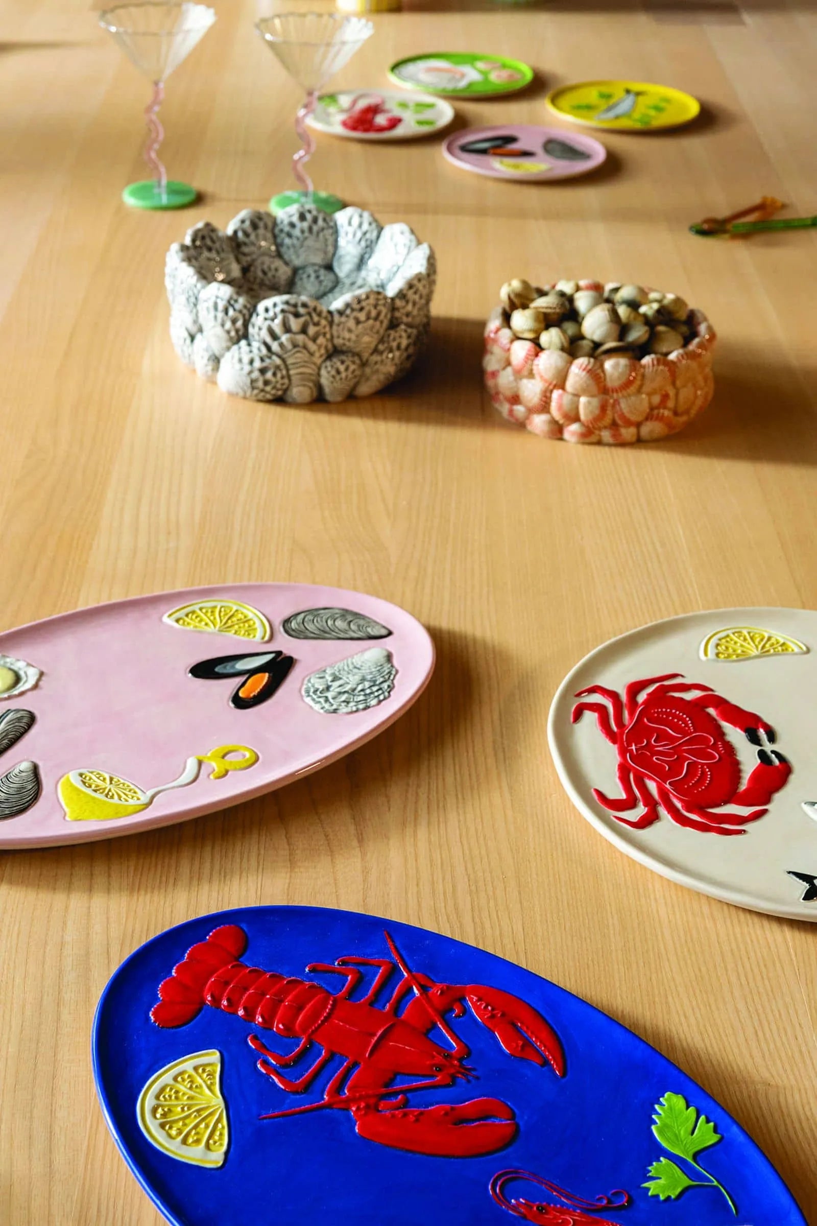 Colorful &klevering ceramic plates featuring hand-painted seafood designs—lobster, crab, oysters, and mussels—bring oceanic charm to a wooden table with shells and two bowls of nuts. Each echoes the &klevering De La Mer Oyster Platter.