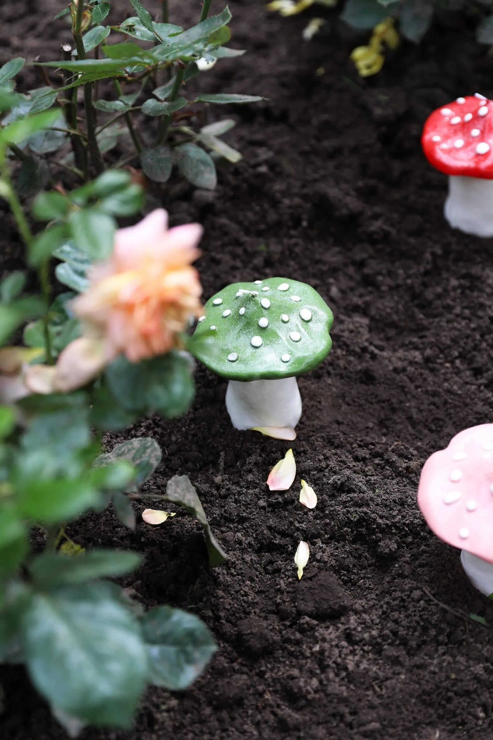 The &klevering Dots Mushroom Candle by &klevering, a green candle with white spots, stands in dark soil near a blurred flower, adding an enchanting touch beside pink and red decorative mushrooms in the background.