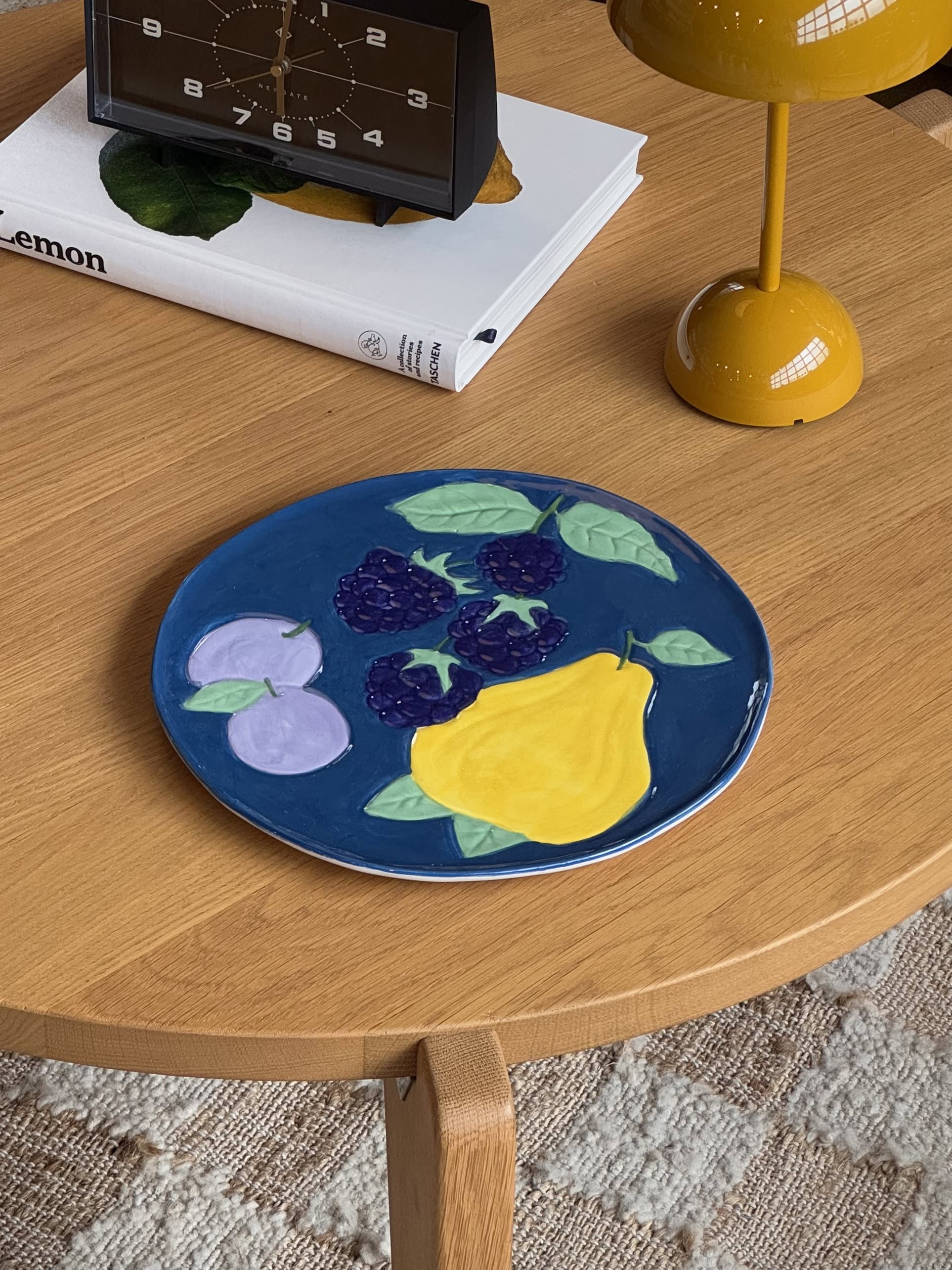 The &klevering Fruitful Blackberry Platter, featuring a vintage fruit motif, sits on a coffee table alongside a yellow lamp, a clock, and Lemon for an inviting table setting.